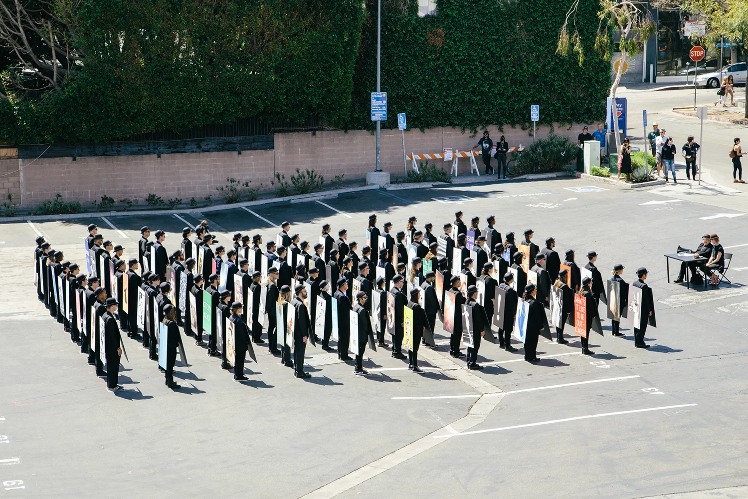 100 Walkers, West Hollywood , a large scale performance piece by Richard Kraft took place on April 18th 2015. One hundred people, each dressed in a dark suit and bowler hat and wearing a sandwich board (with a unique image on both front and back) walked pre-determined routes through the city. The piece began with all one hundred walkers forming a grid in the El Tovar parking lot adjacent to West Hollywood Park. Once all one hundred walkers were in place, the maestra of ceremonies Marjorie Perloff began calling "start phrases" to release one walker at a time at approximately five second intervals. Once the walkers dispersed, they could be seen all over West Hollywood, particularly along all the main thoroughfares: Sunset, Santa Monica, Melrose, Beverly, San Vicente, Doheny, La Cienega, Crescent Heights and Fairfax. During the piece performers remained silent. Interaction with inquisitive passers by was confined to the handing out of a business card. The walk ended when all walkers returned to their starting point at the El Tovar parking lot, standing for one minute to finish their performance. For more images and further information please visit: www.onehundredwalkers.com Photo by Tony Coehlo
