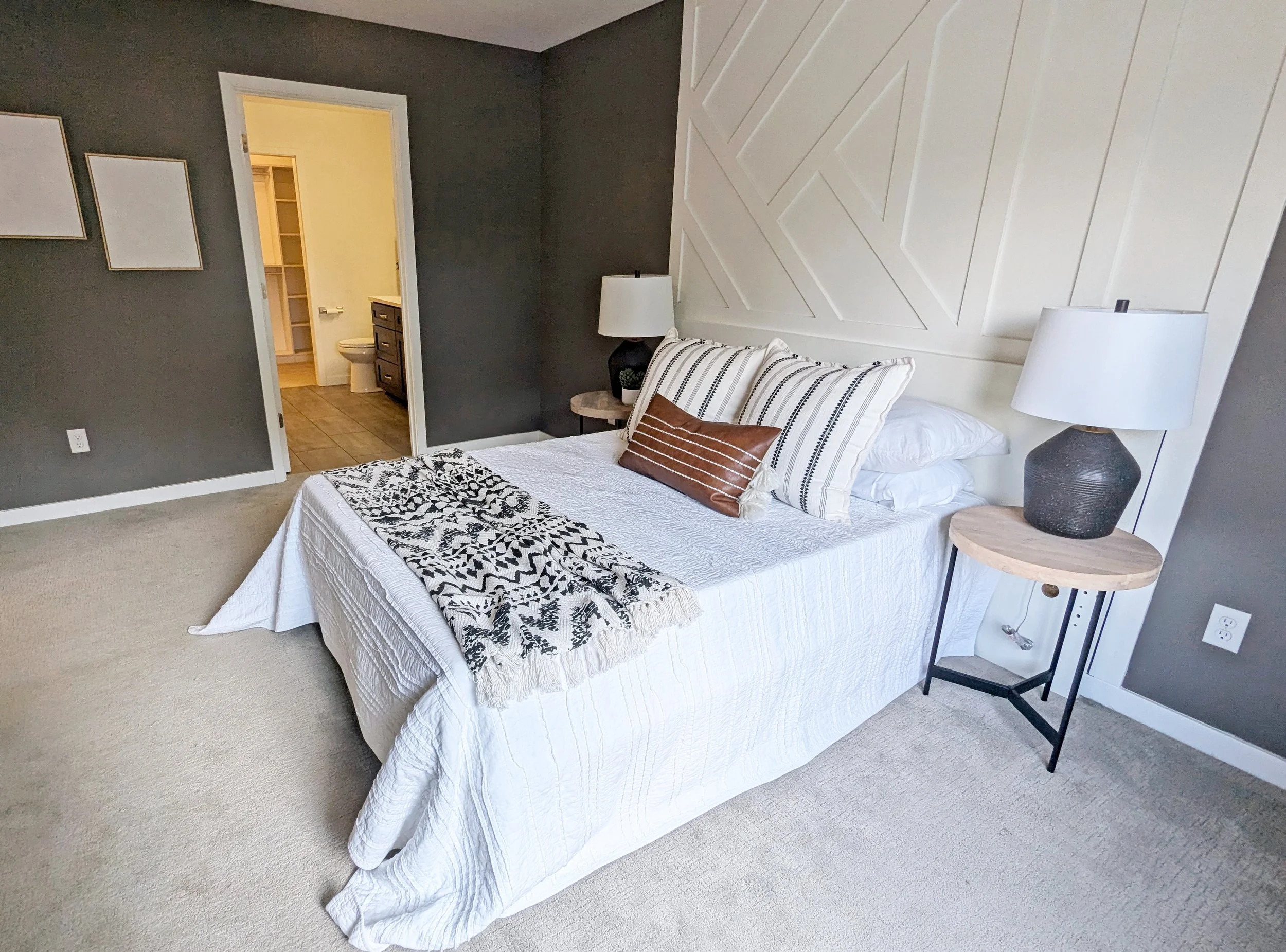 A bedroom with a white bedspread, decorative pillows, black and white throw blanket, side table with a plant, a table lamp, and a dark gray accent wall with three abstract framed art pieces; an open doorway leads to a bathroom with a toilet and storage shelves.