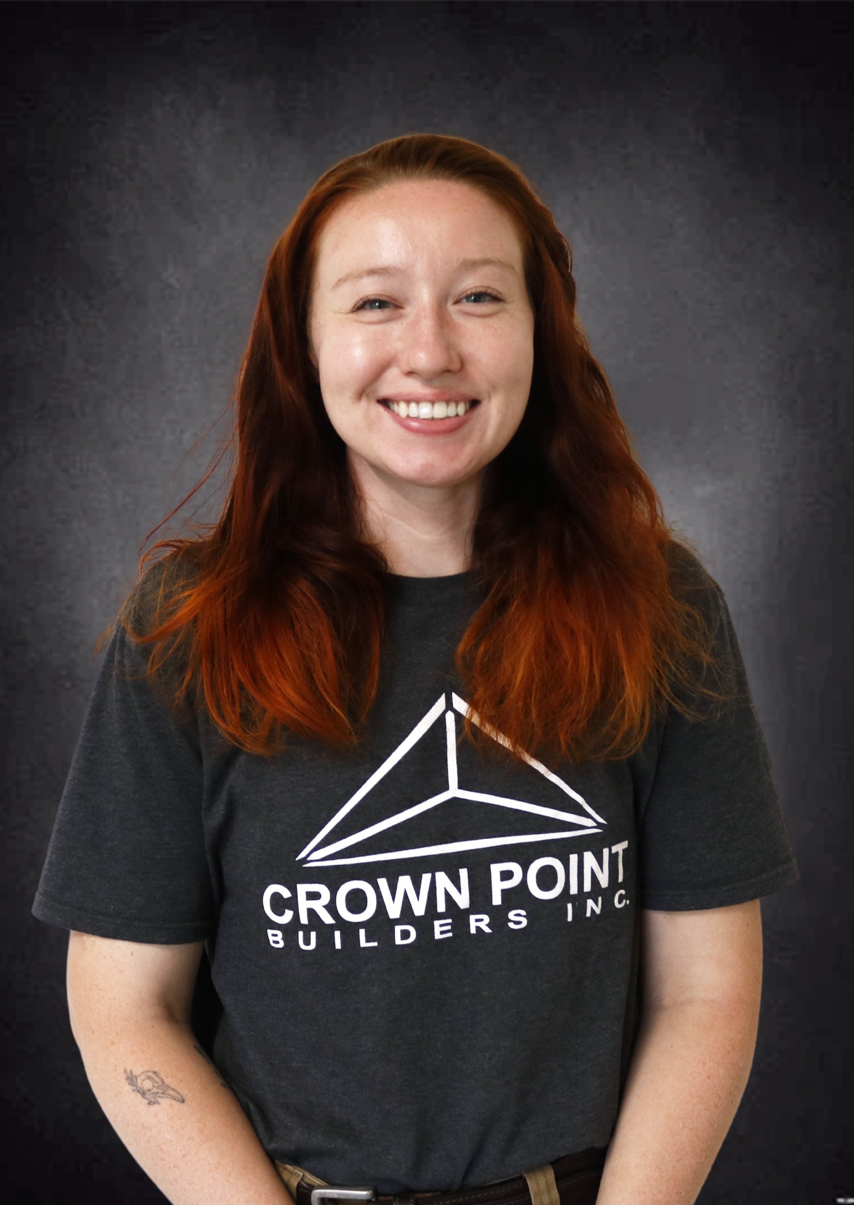 A smiling woman with red hair, wearing a dark gray T-shirt with the logo and name 'Crown Point Builders Inc.' on it, posing in front of a dark textured background.