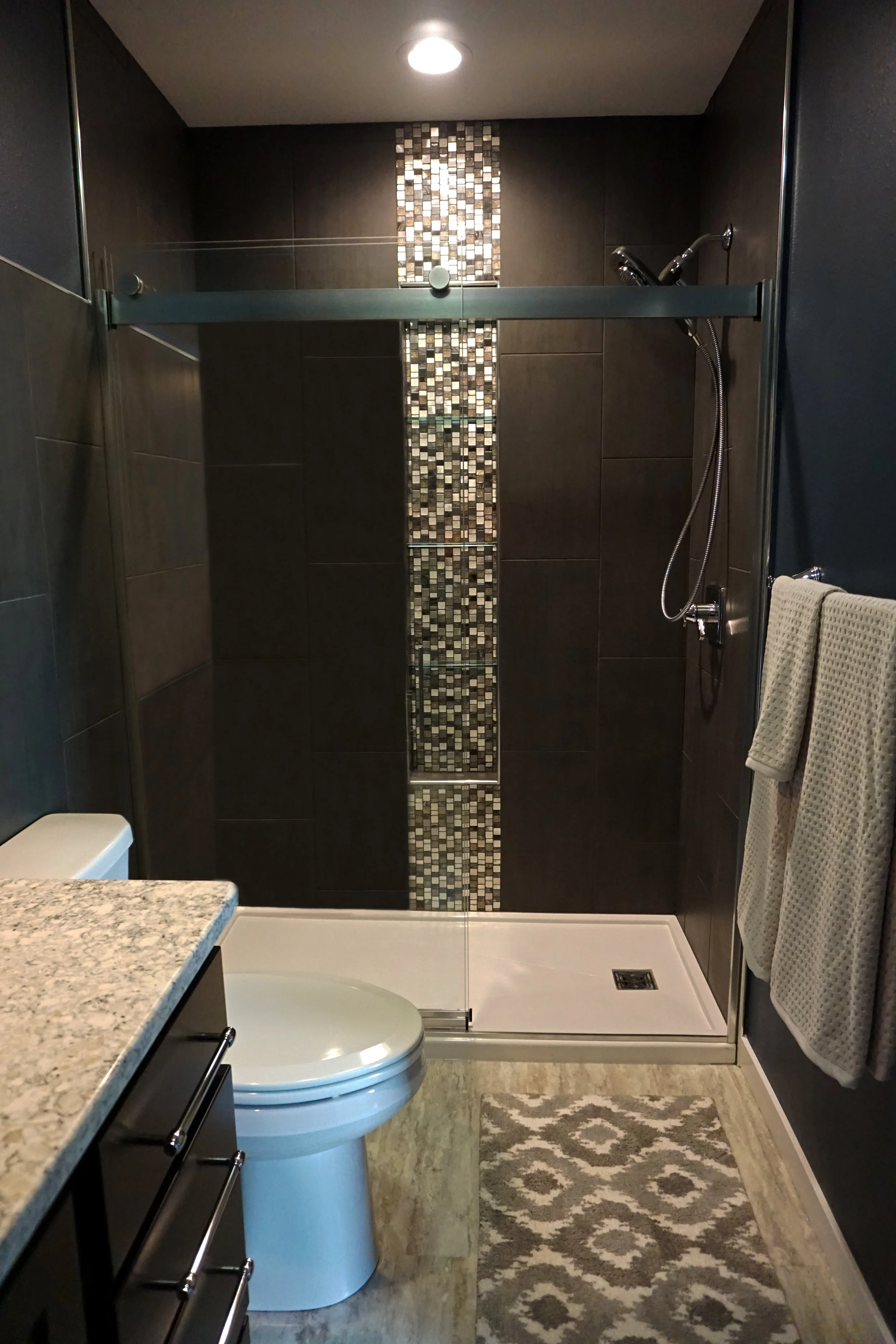 A modern bathroom with a walk-in shower that has dark brown tiles, a decorative vertical strip of small mosaic tiles, a chrome showerhead, and a glass door. To the right, there are two beige towels hanging on a towel rack. In the foreground, there is a bathroom vanity with a granite countertop, a toilet, and a patterned rug on the floor.