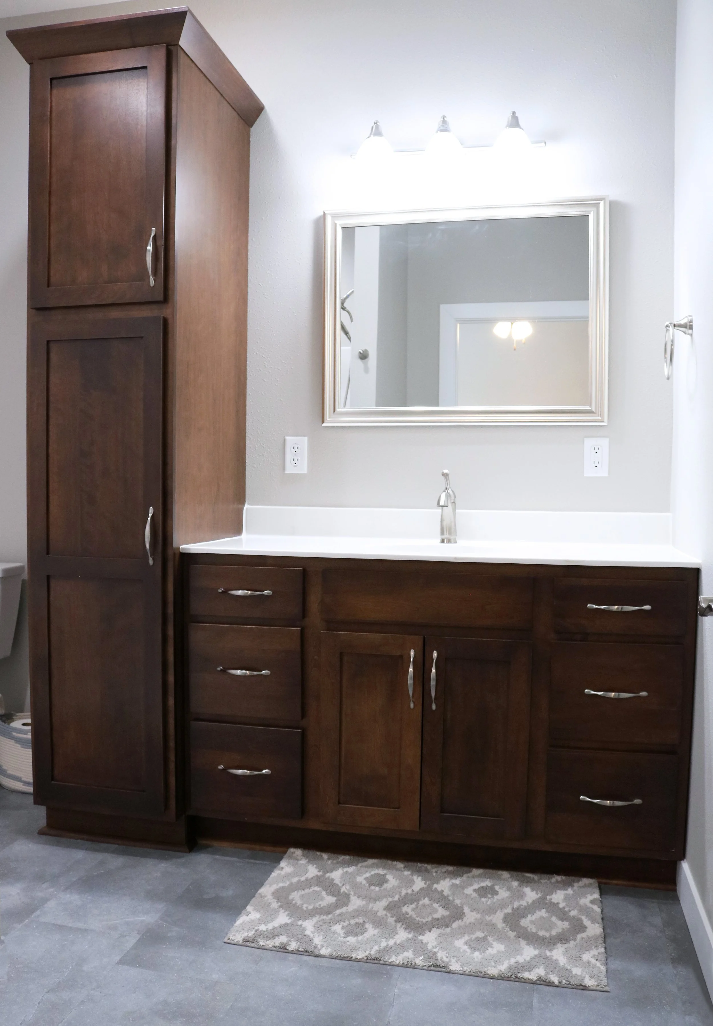 Bathroom vanity with dark wood cabinets, a white countertop, a framed mirror, and a three-light fixture, with a small beige patterned rug on gray tiled floor.