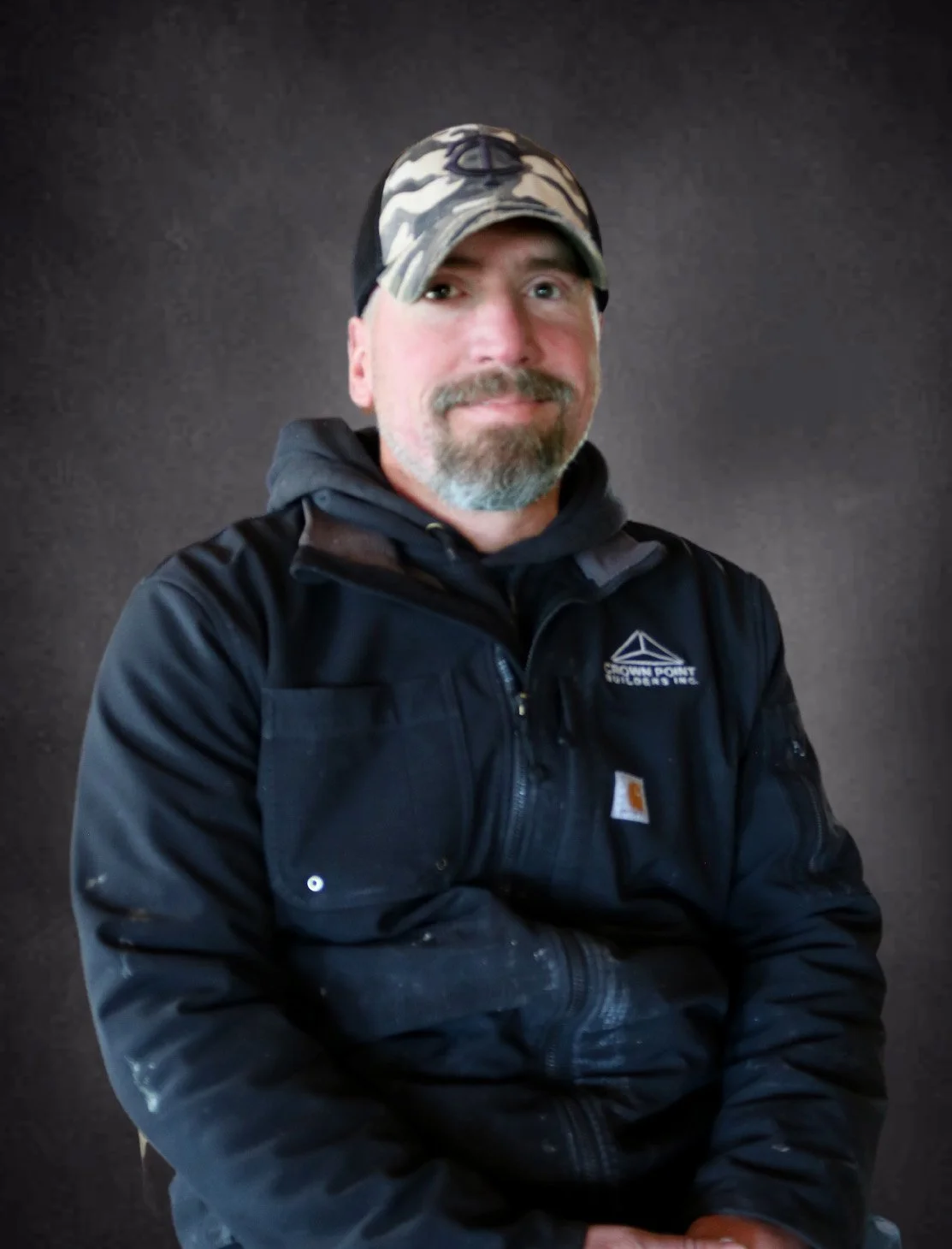 A man wearing a camouflage baseball cap, black Carhartt jacket, and gray hoodie, sitting against a dark background.