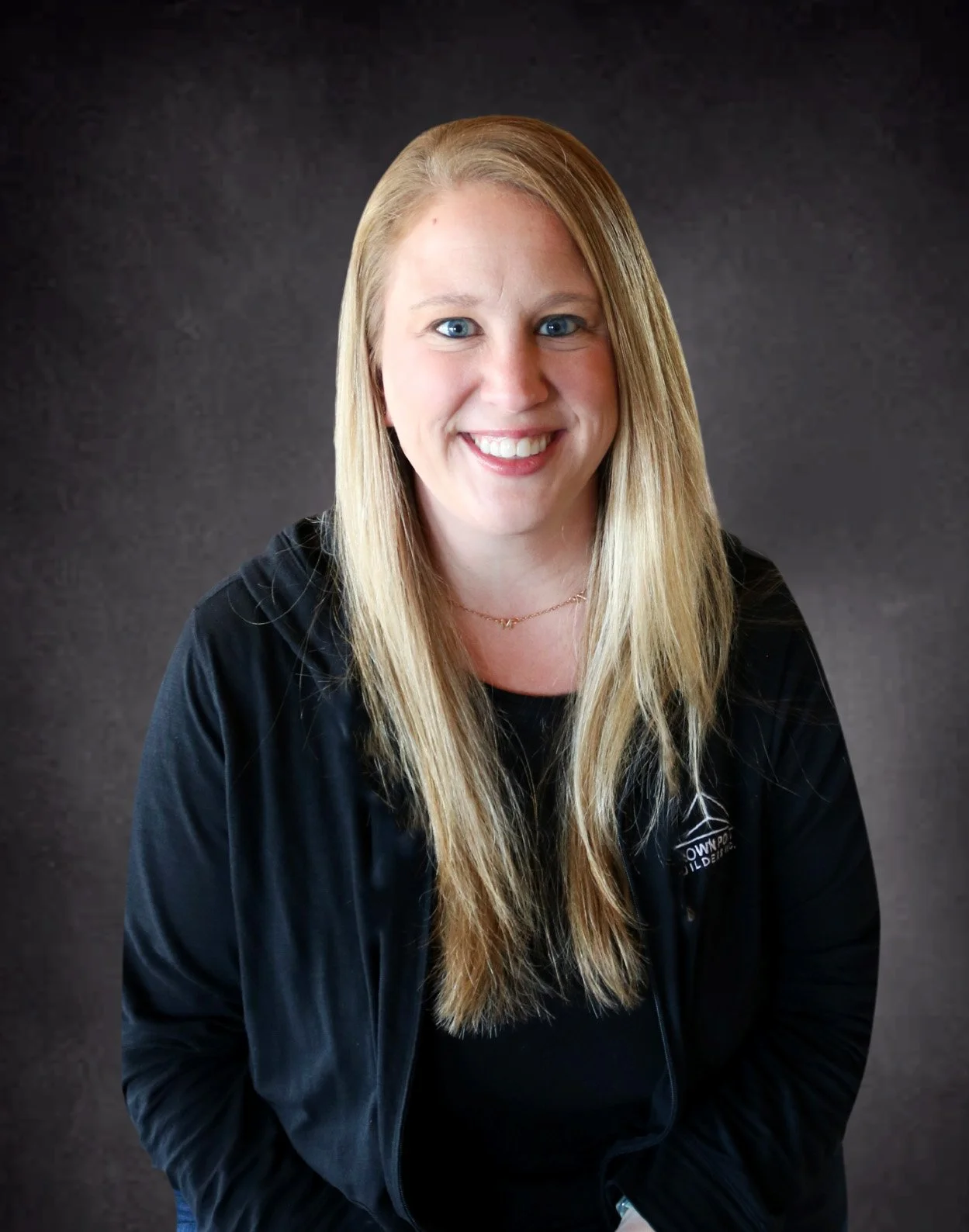 Portrait of a smiling woman with long blonde hair, wearing a black jacket and a black shirt, against a dark background.