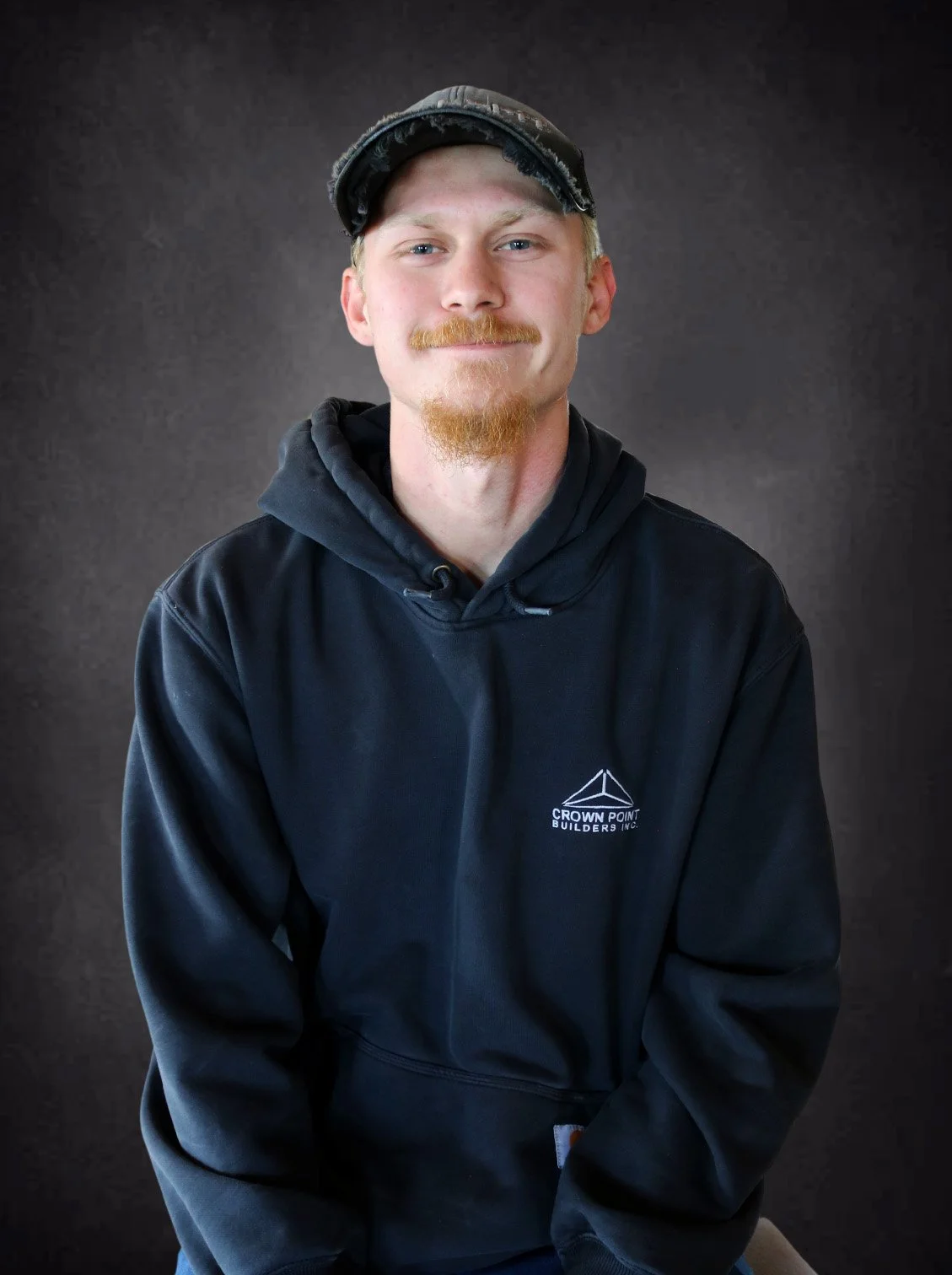 A young man with blonde hair and a goatee, wearing a black hoodie and a camo cap, smiling and sitting against a dark background.