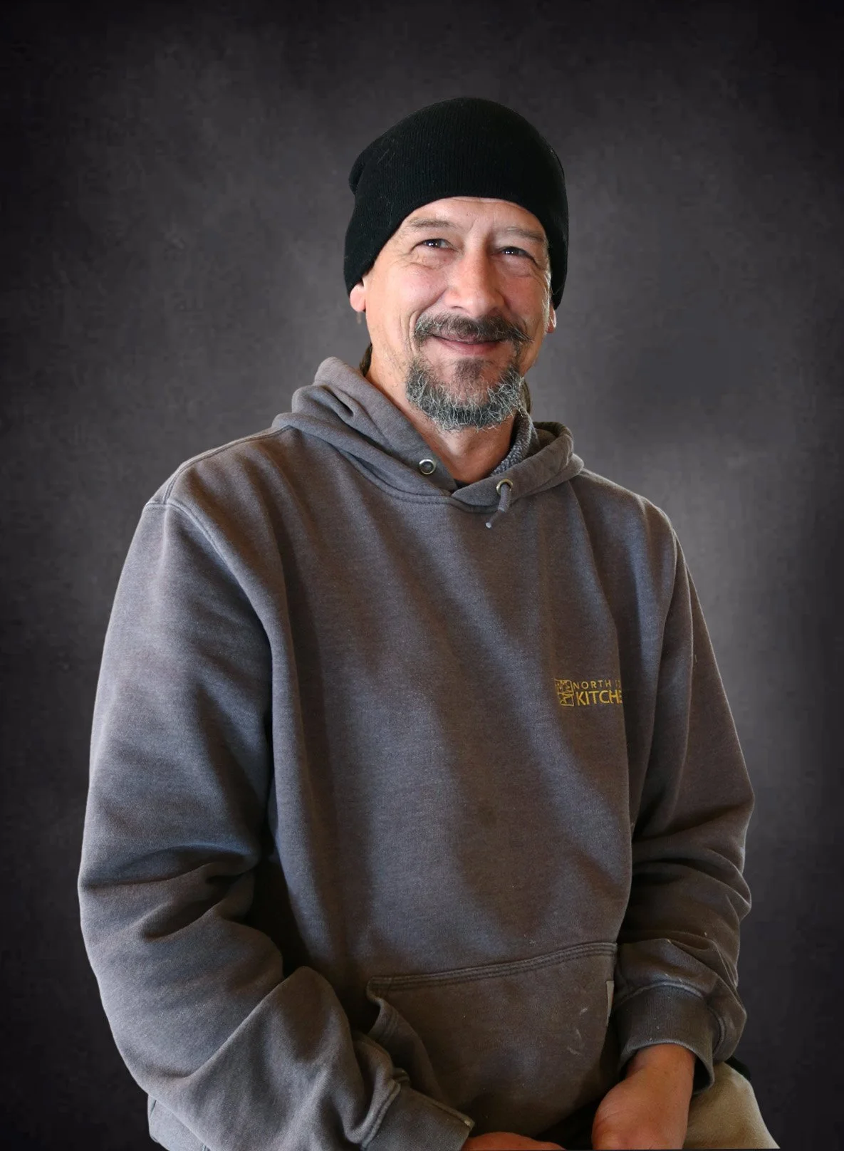 A middle-aged man with a goatee, wearing a black beanie, gray hoodie with a small yellow logo, and smiling in front of a dark textured background.