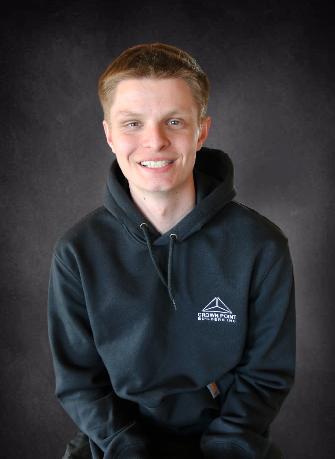 Young man with short light brown hair smiling, wearing a black hoodie with a logo and text, against a dark background.