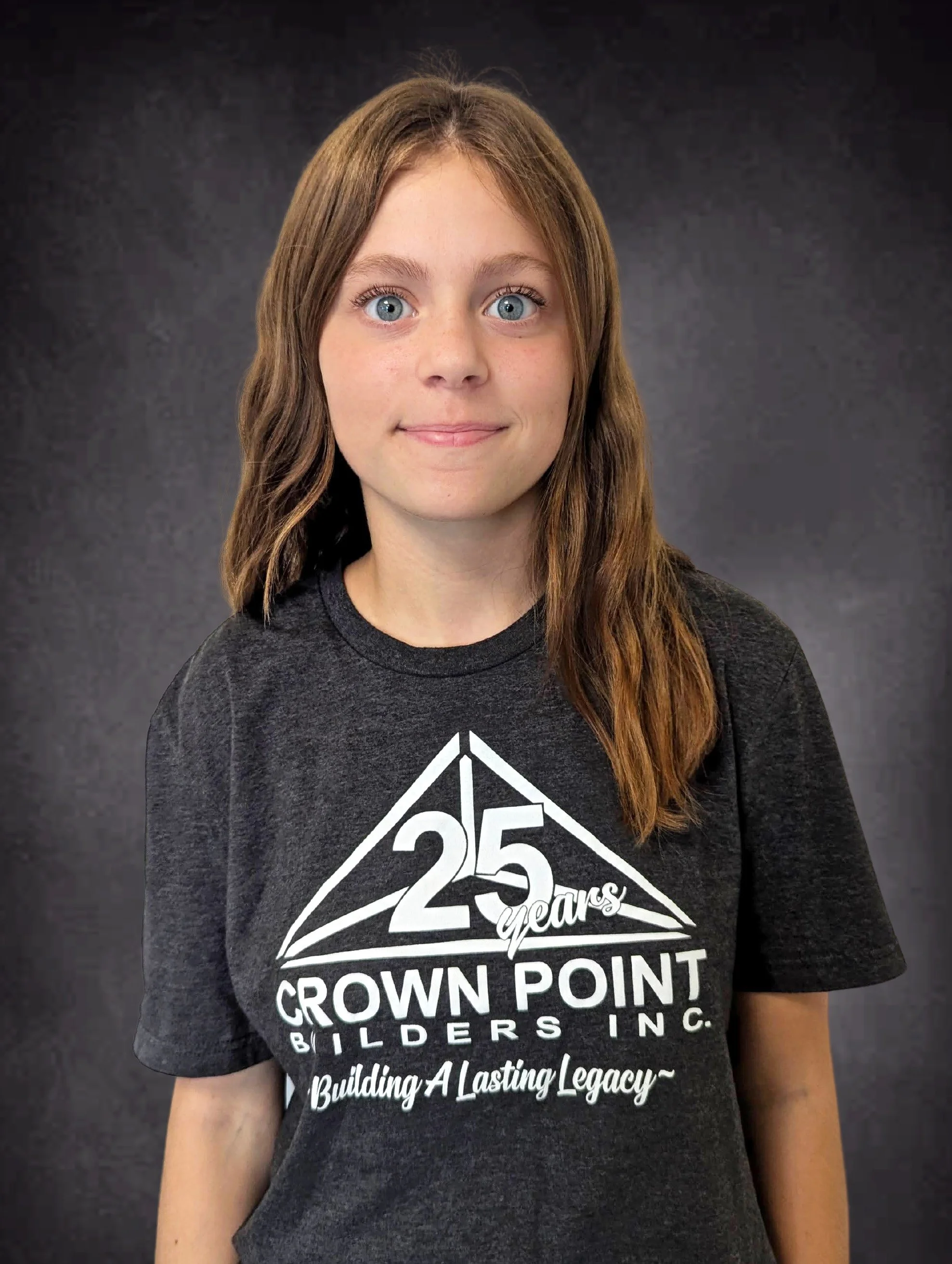 A young woman with blue eyes and brown hair standing in front of a dark background, wearing a black T-shirt celebrating 25 years of Crown Point Builders Inc.