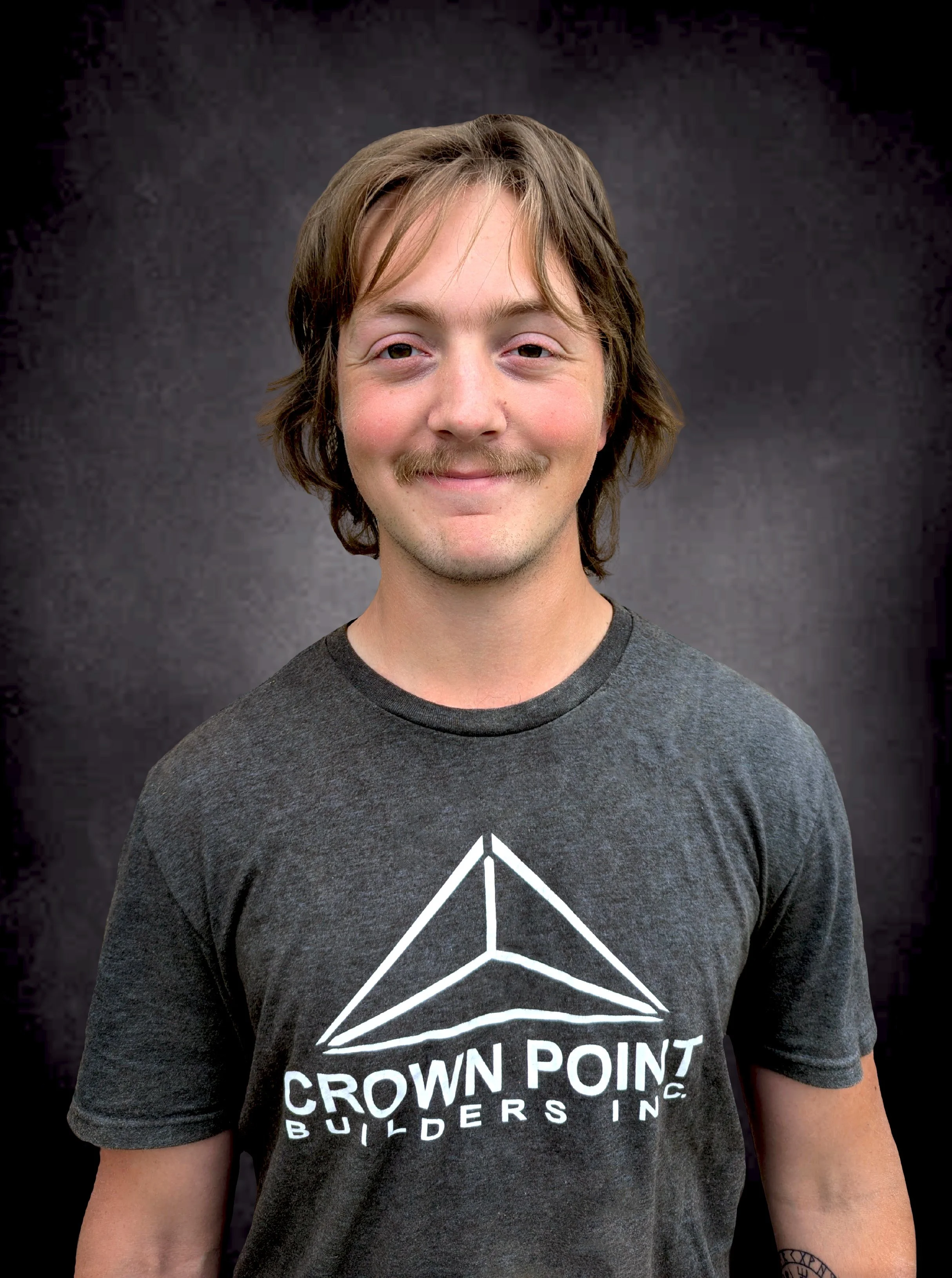 A young man with shoulder-length light brown hair, a mustache, and a goatee, wearing a dark gray T-shirt with a white geometric logo and text reading 'Crown Point Builders Inc.' standing against a dark, textured background.