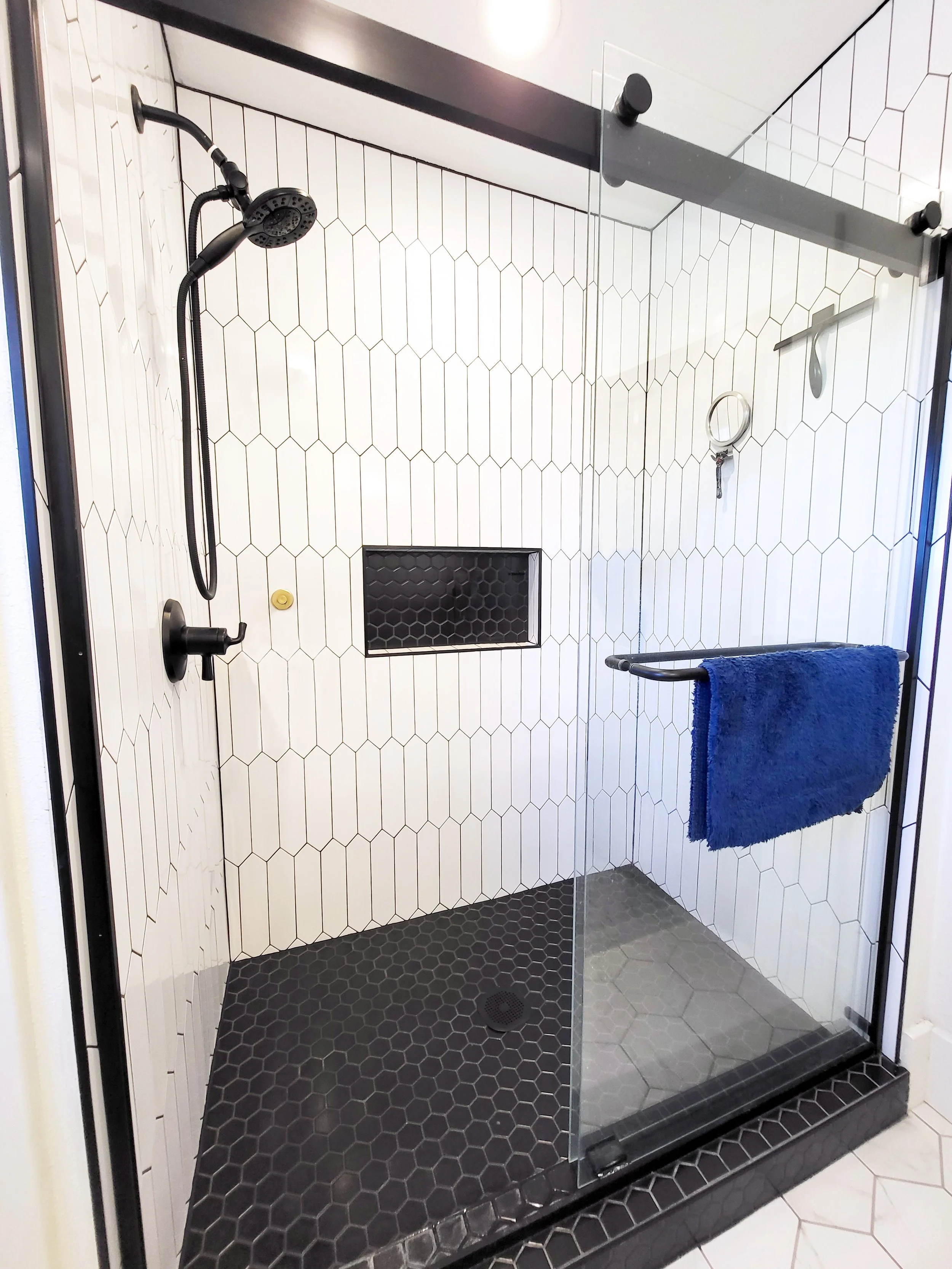 A walk-in shower with black floor tiles, white wall tiles with black grout lines, a black shower head, a small black drain, a glass door, a black grab bar with a blue towel, and a honeycomb shower niche.