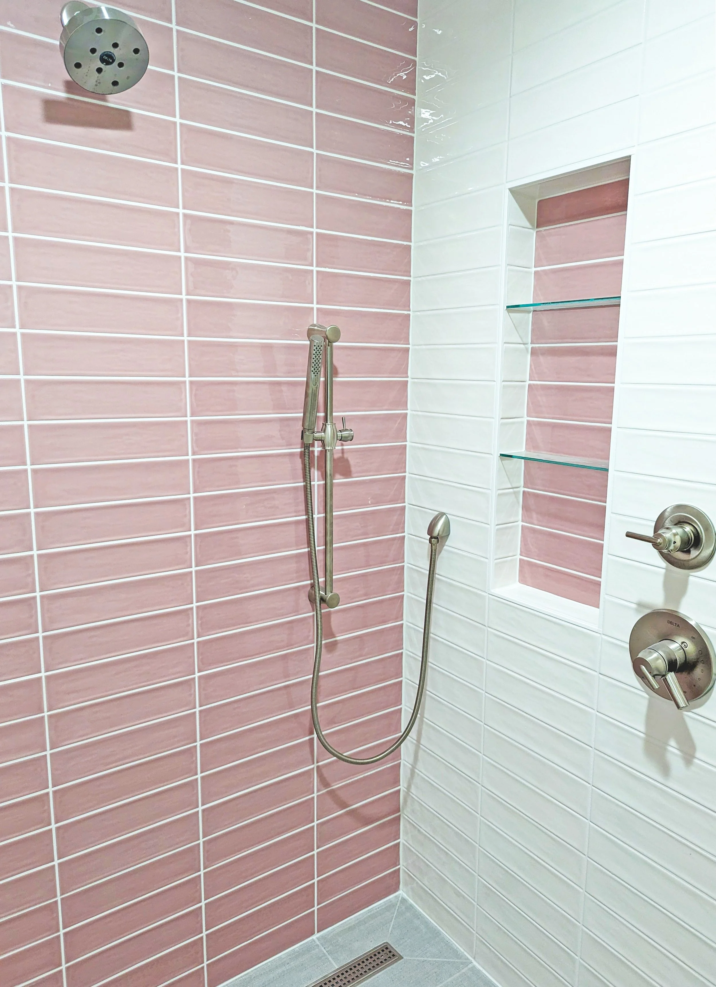 A shower area with pink and white tiled walls, a showerhead, a handheld shower, and a built-in shelf with glass shelves.