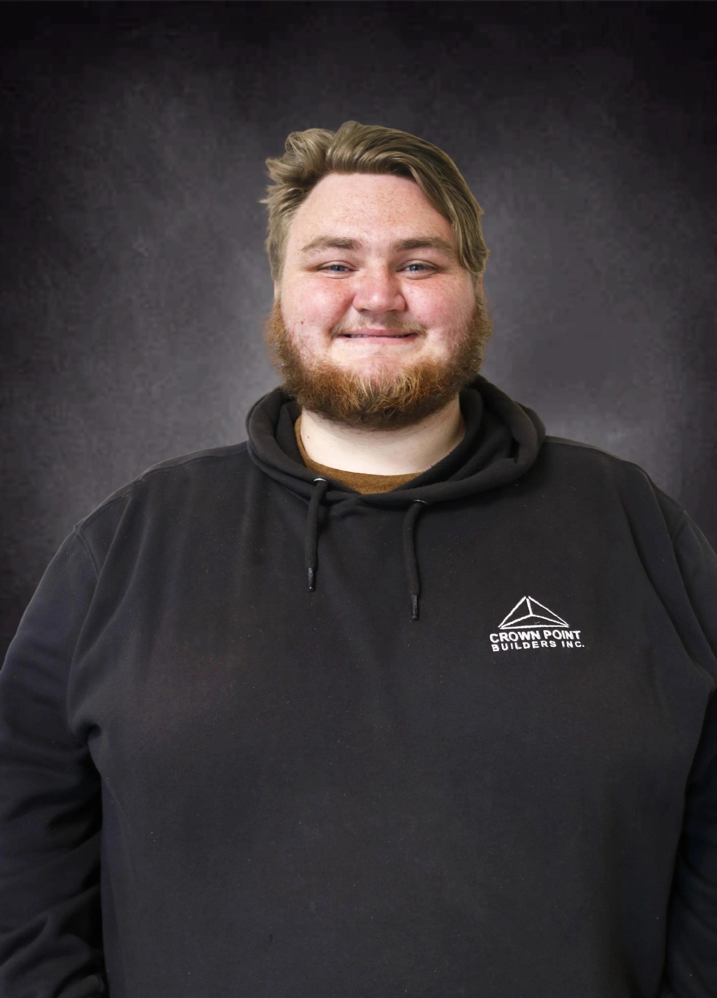 A young man with a beard and short, wavy hair, smiling, wearing a black hoodie with a logo on the chest that says 'Crown Point Builders Inc.' against a dark background.