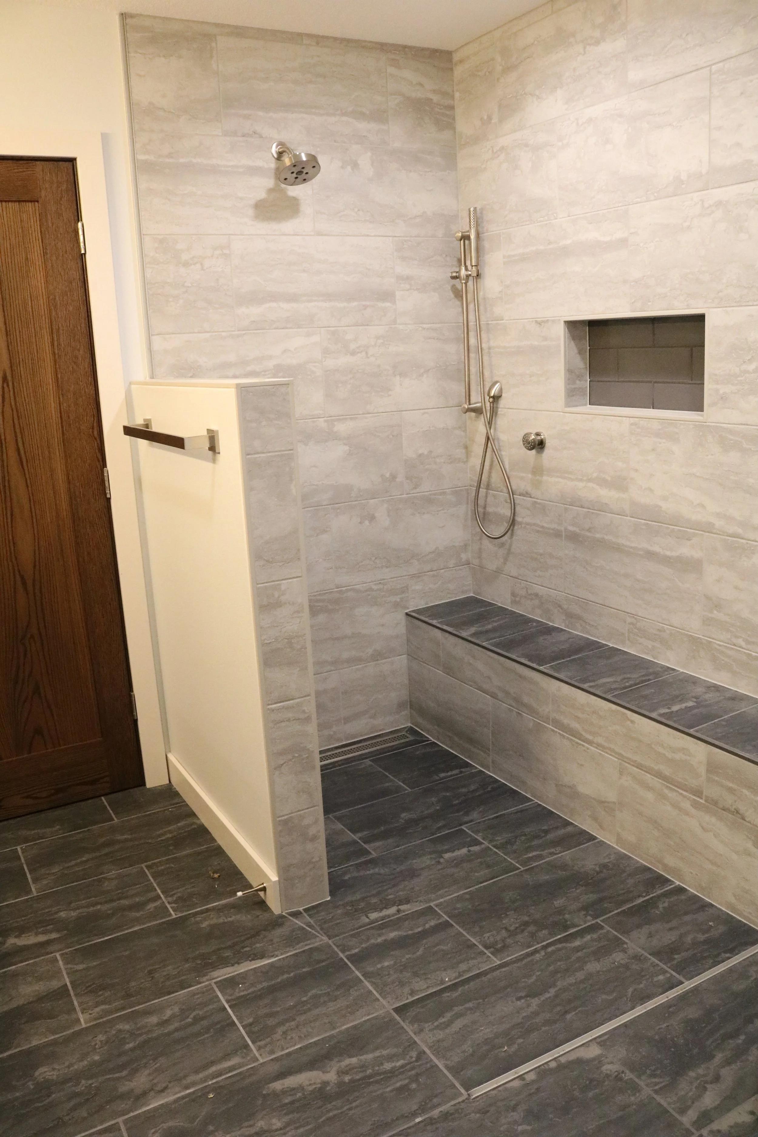 A modern walk-in shower with gray textured wall tiles, a showerhead, a hand-held shower wand, a built-in niche for toiletries, a bench with darker gray tiles, and a floor drain.