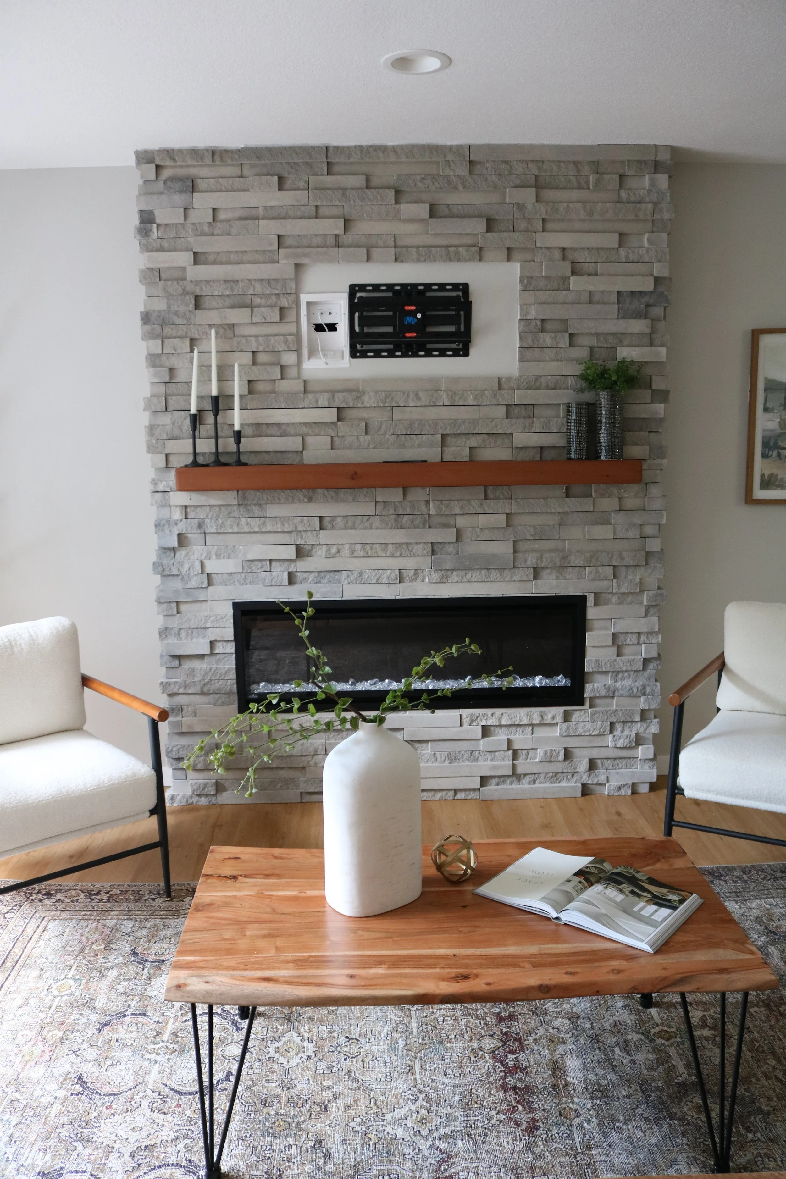 Living room with stone fireplace, wooden mantel, white chairs, a wooden coffee table with a tall white vase and an open magazine, decorative items, and potted plants.