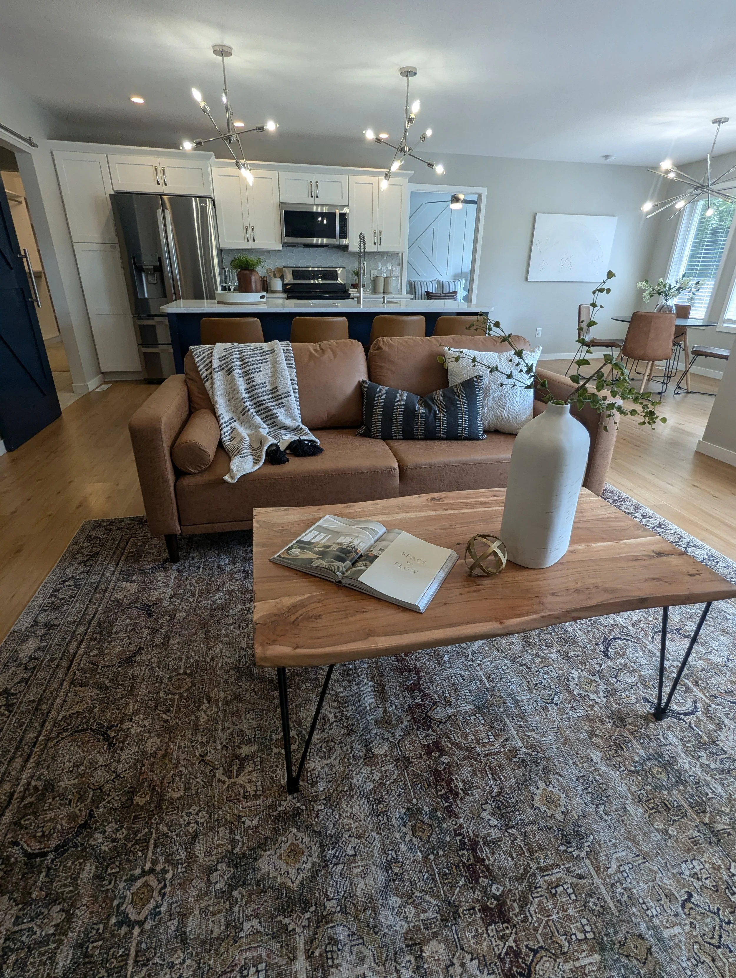 Living room with a beige sofa, a wooden coffee table, a decorative horse sculpture, and an open book. The background features a kitchen with white cabinets, stainless steel appliances, a gray hexagonal tile backsplash, and a dining area with a round table and four chairs near a window with blinds. The room has hardwood flooring and modern lighting fixtures.