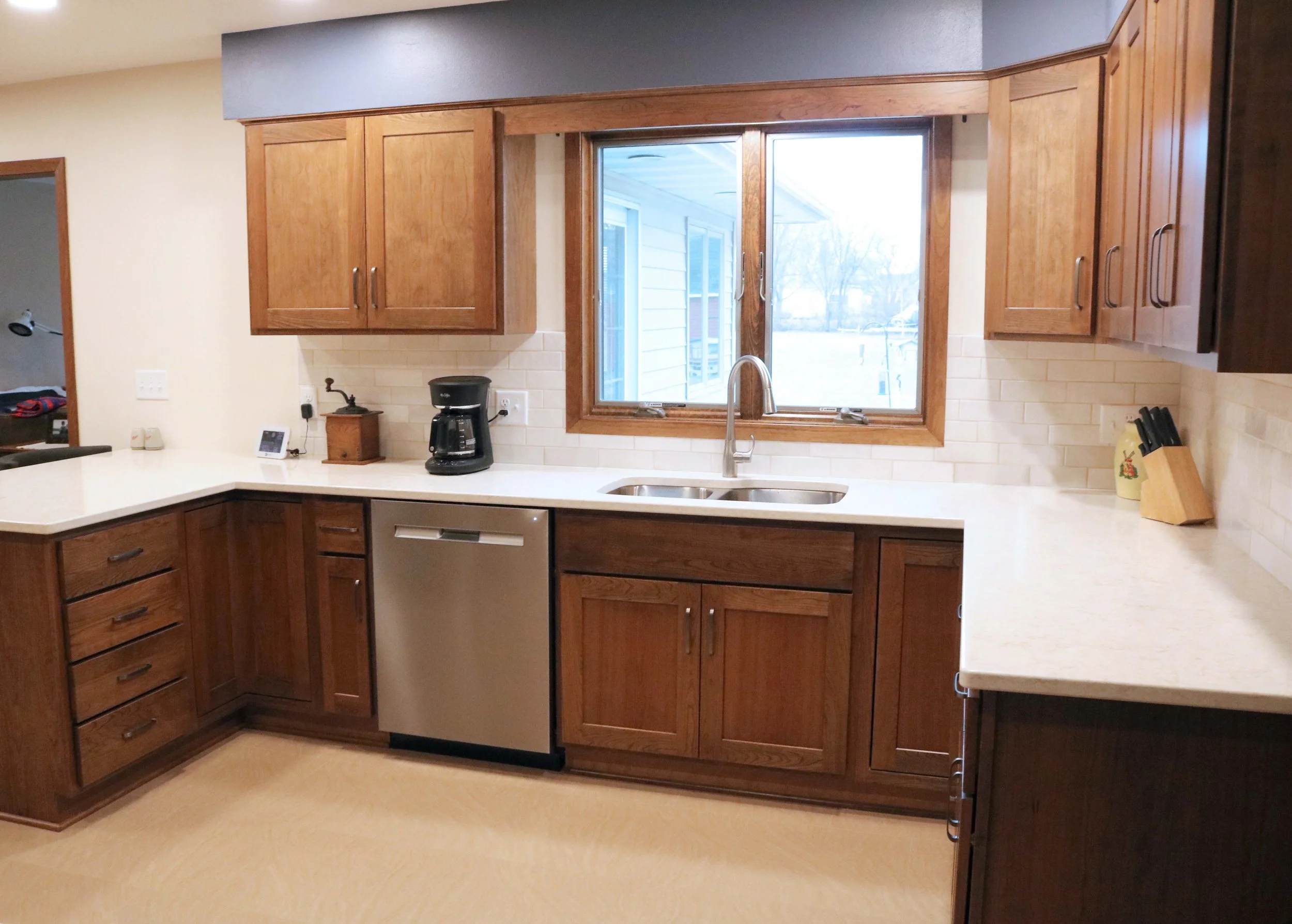 Modern kitchen with wooden cabinets, stainless steel dishwasher, coffee maker, double sink, knife block, and white countertops.