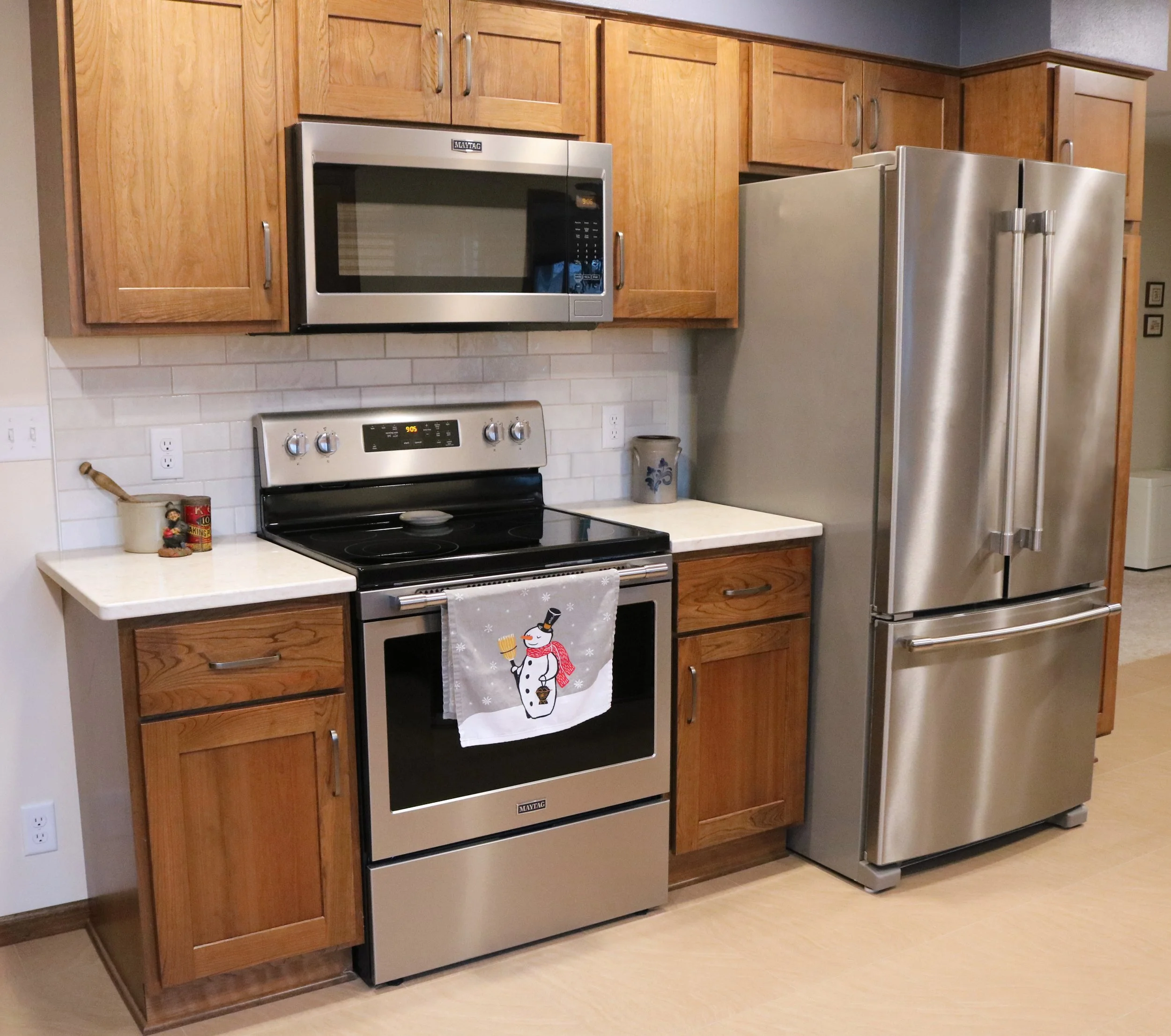 Kitchen with wooden cabinets, stainless steel microwave and refrigerator, electric stove, and countertop with a decorative snowman towel hanging on the oven handle.