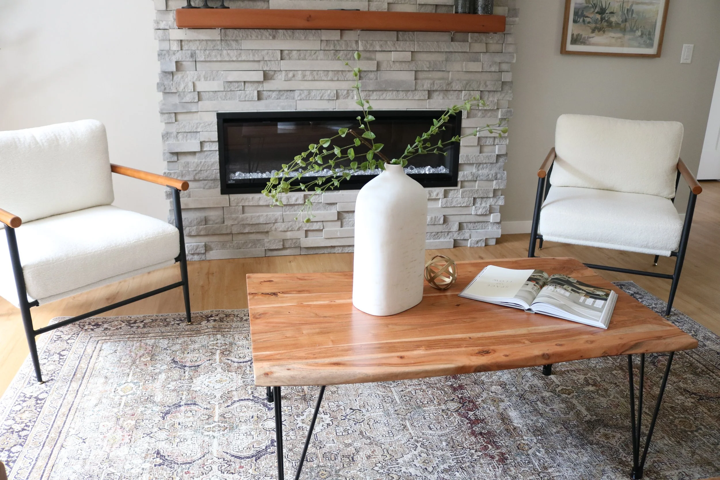 Living room with a stone fireplace, two white armchairs with wooden arms, a wooden coffee table with a white vase and green plant, an open magazine, a patterned area rug, a wall art painting, and hardwood flooring.