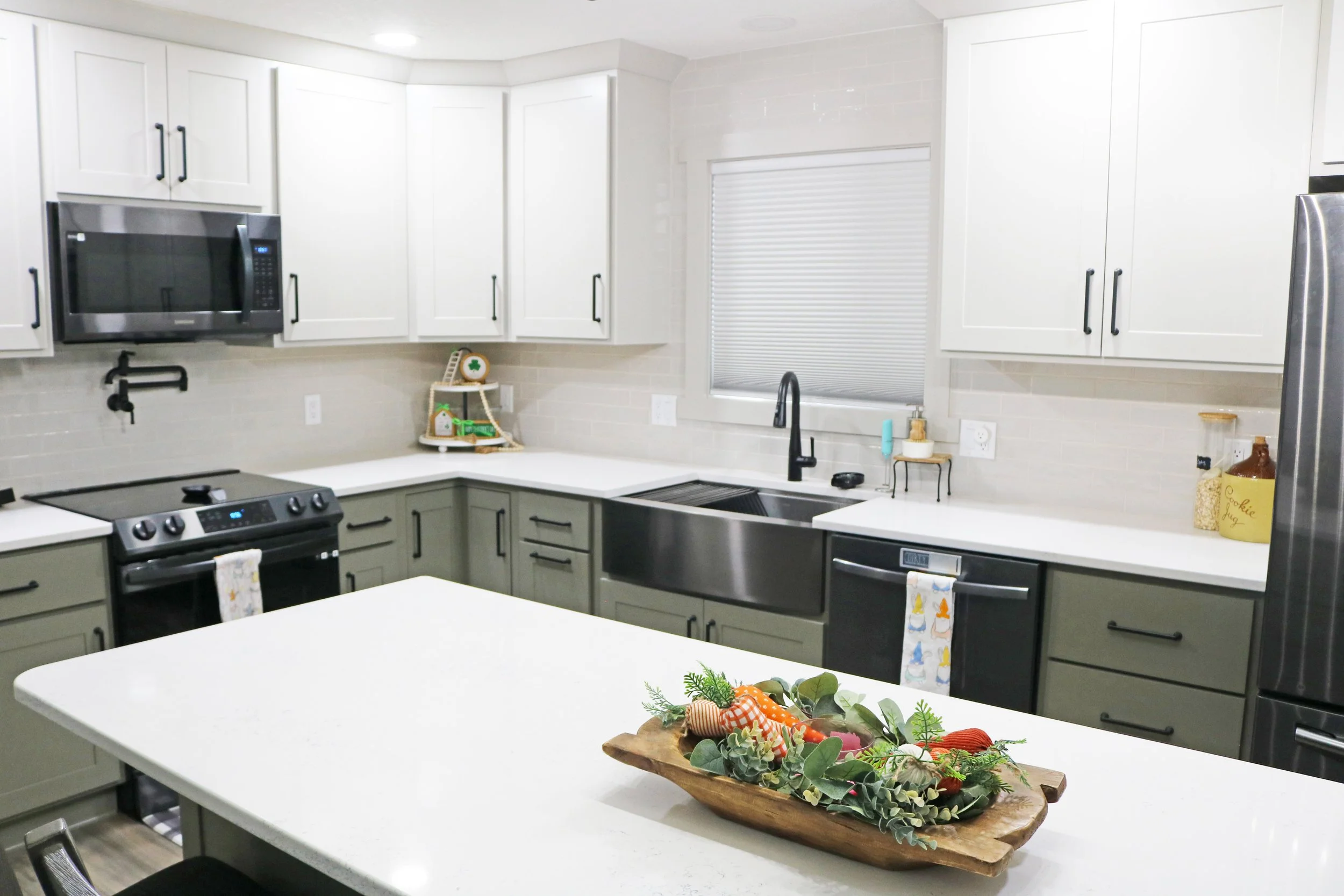 Modern kitchen with white countertops, gray lower cabinets, white upper cabinets, stainless steel microwave and refrigerator, black sink, window with closed blinds, decorative fall-themed arrangement on island.