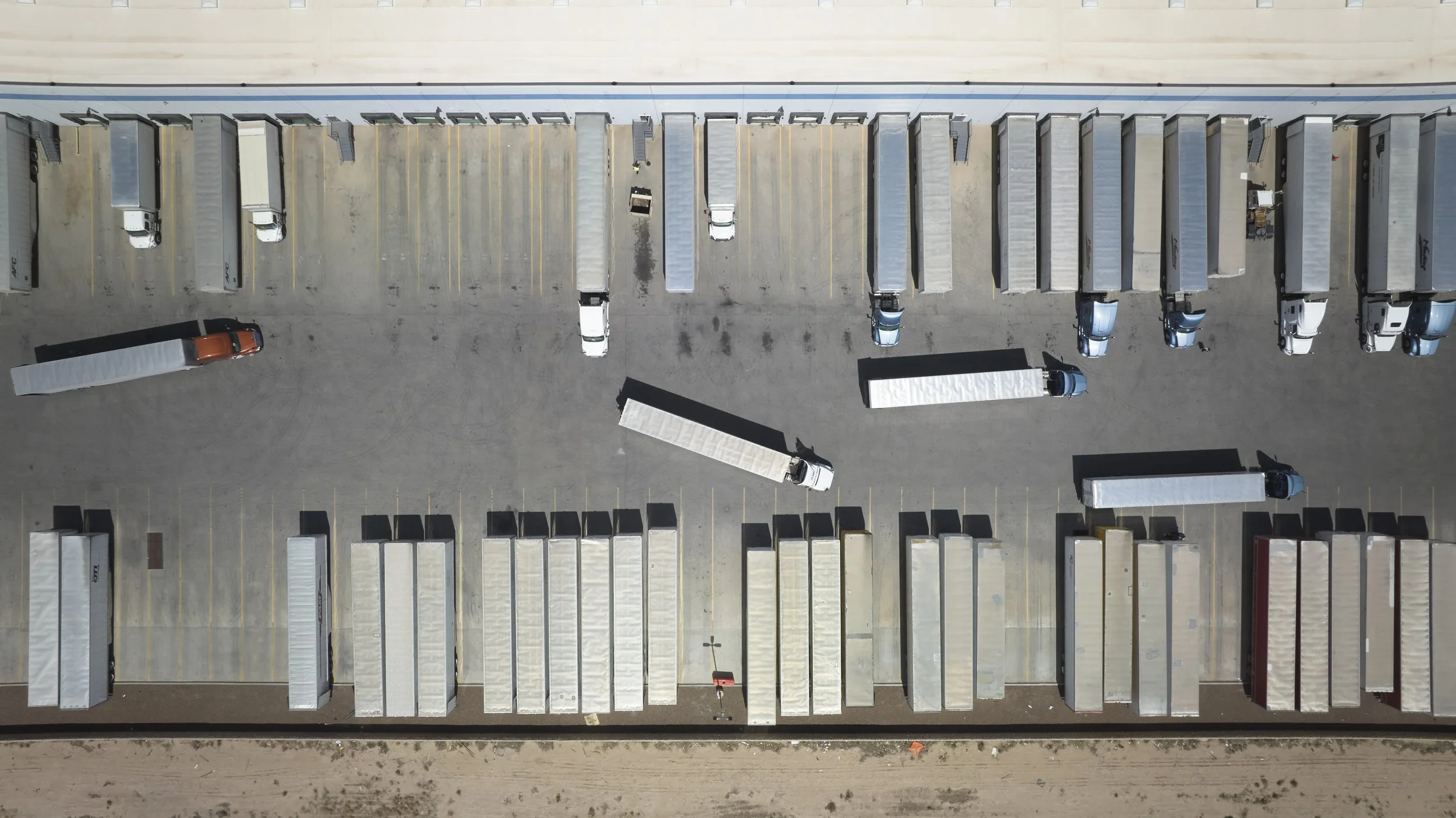 An aerial view of a parking lot filled with semi-truck trailers and trucks, with some trailers parked along the top and bottom edges and a few trucks parked in between.