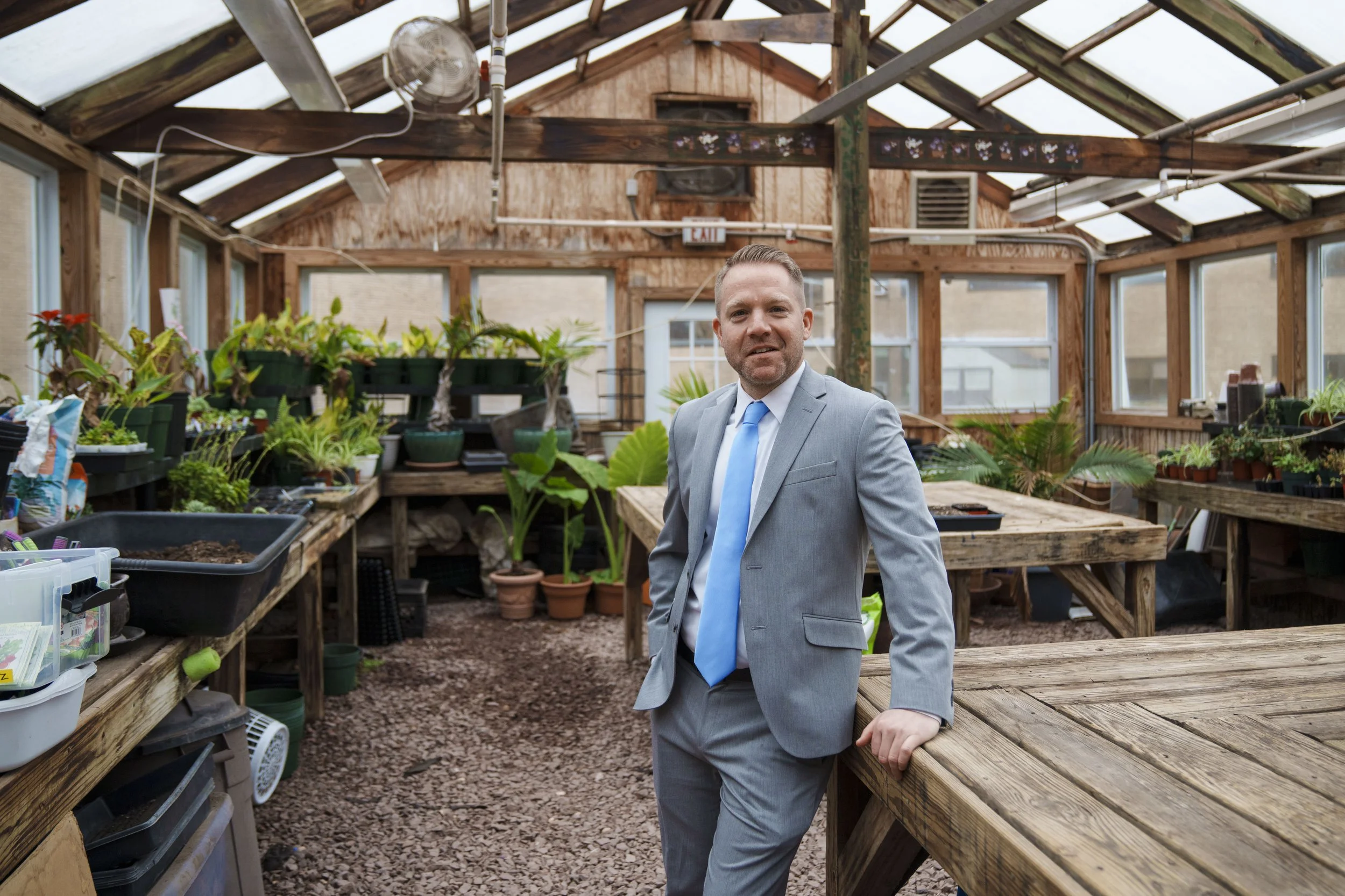 A man in a gray suit with a blue tie standing inside a greenhouse with wooden frames, surrounded by various plants and gardening supplies.