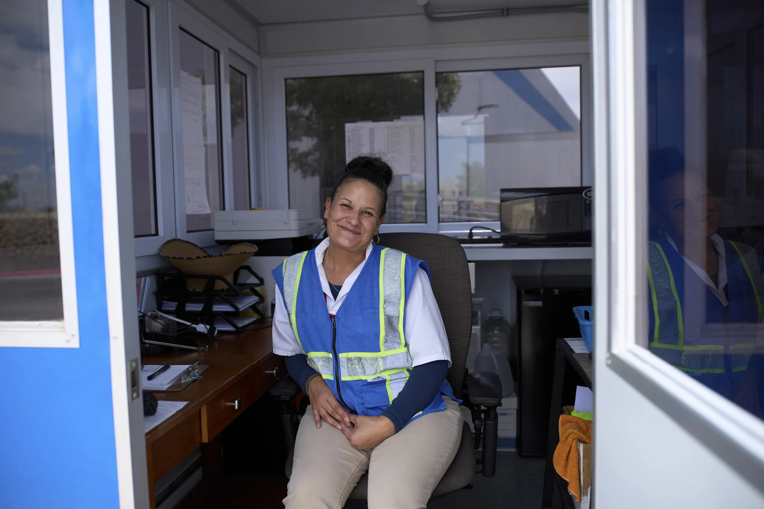 A smiling woman in a blue safety vest sitting inside a small office booth, with office supplies and a microwave visible behind her.