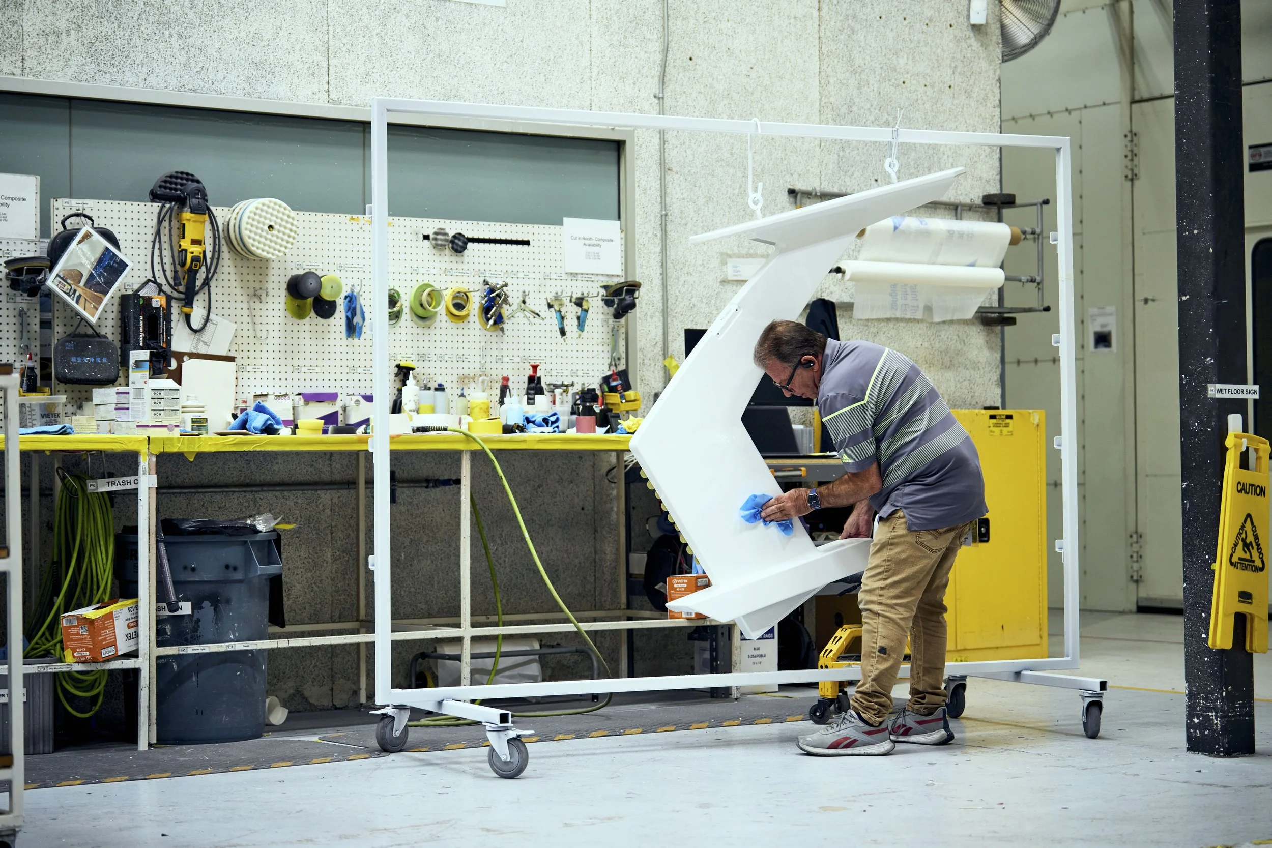 A man in a workshop or manufacturing environment cleaning a white, mounted piece of equipment or furniture with a blue cloth. The area has tools and supplies on a pegboard and a workbench in the background, and signs indicating safety precautions.