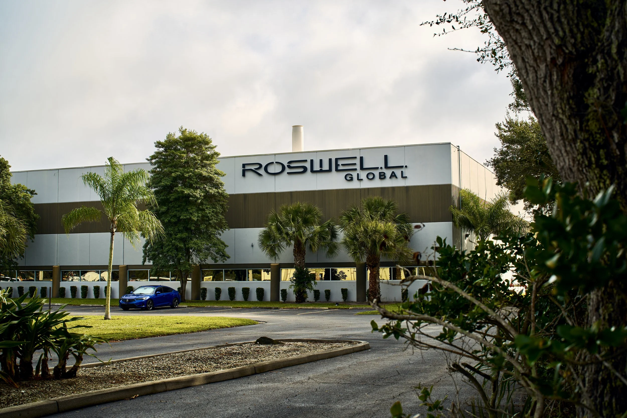 Exterior of a building with a sign that reads "Roswell Global," surrounded by trees and a parking lot with a blue car.