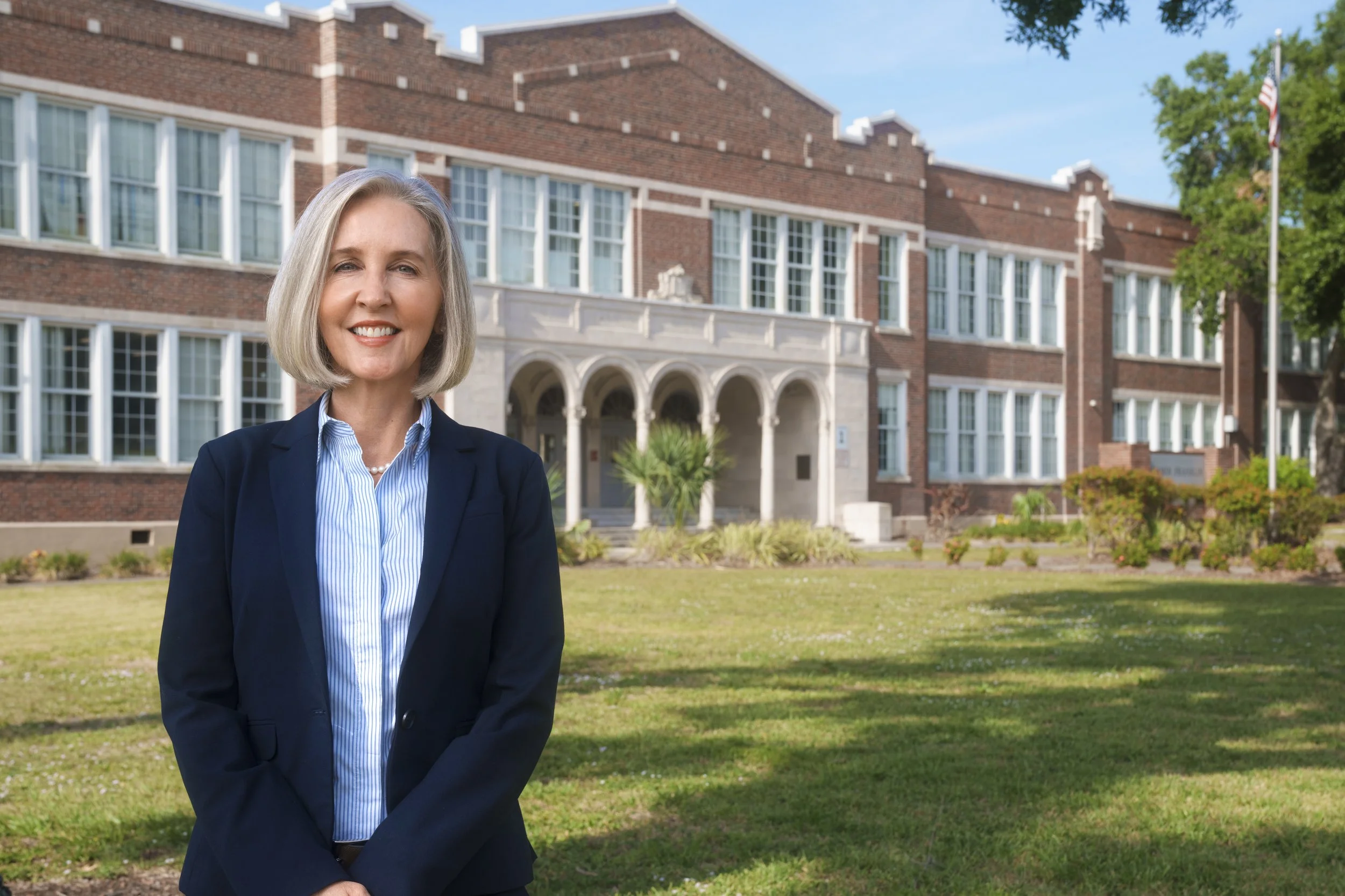 A smiling woman with short gray hair, wearing a dark blazer and light blue shirt, standing outside in front of a college or university building on a sunny day.