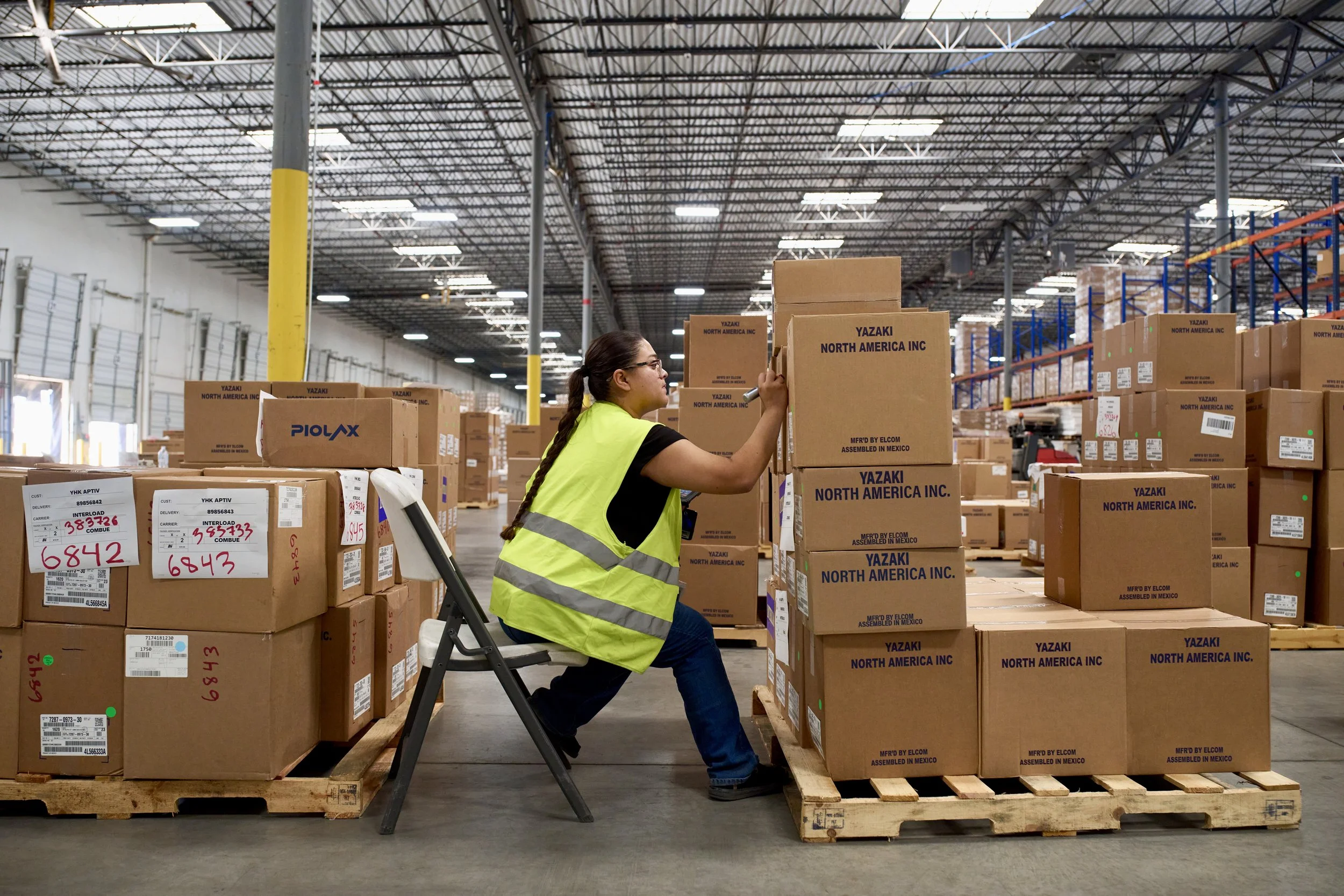 A woman in a yellow safety vest sitting on a chair and stacking cardboard boxes in a warehouse with high ceilings and metal shelving.