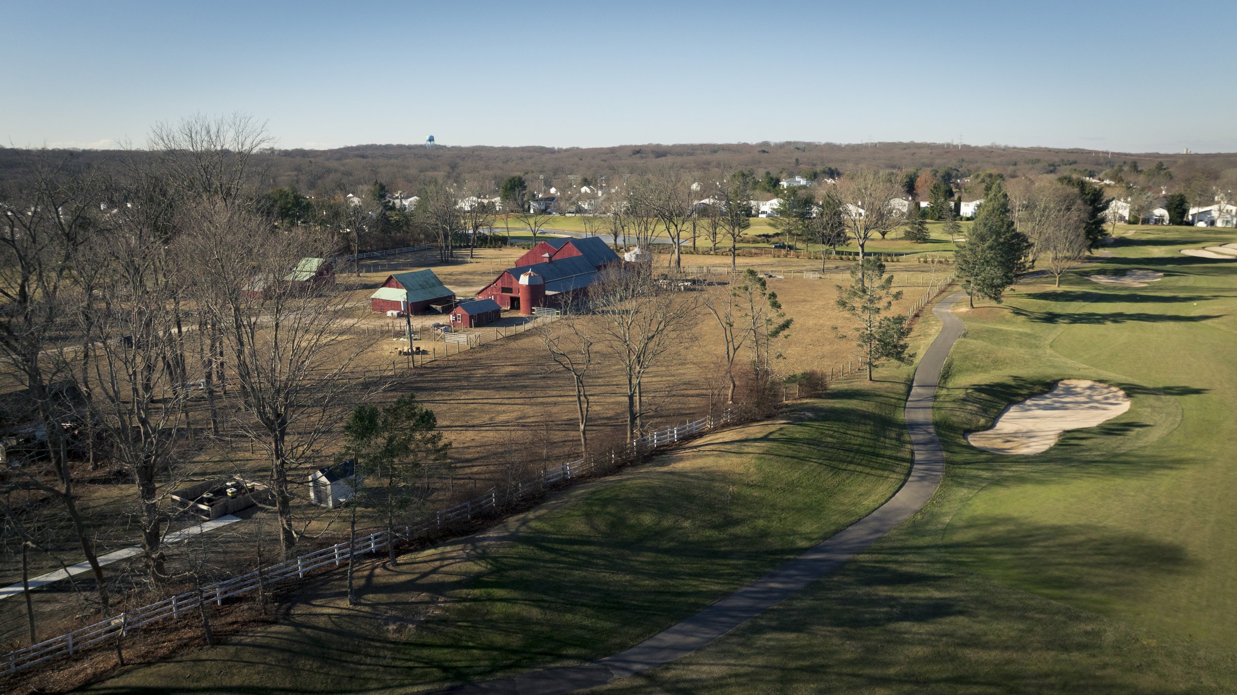 Aerial view of a golf course with a green and sand traps, a pathway, and a red barn among bare trees, with residential houses in the background under a clear blue sky.