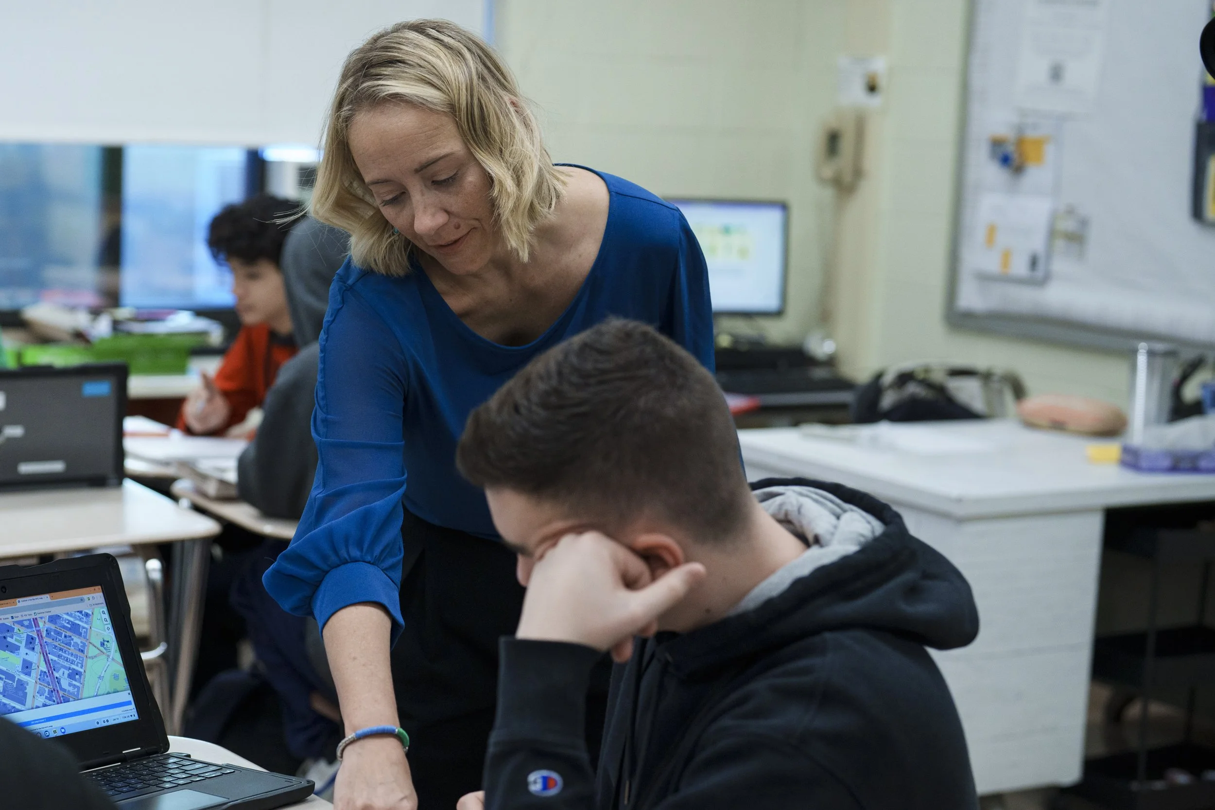 A teacher assists a student in a classroom with other students working at their desks, some using laptops and computers.