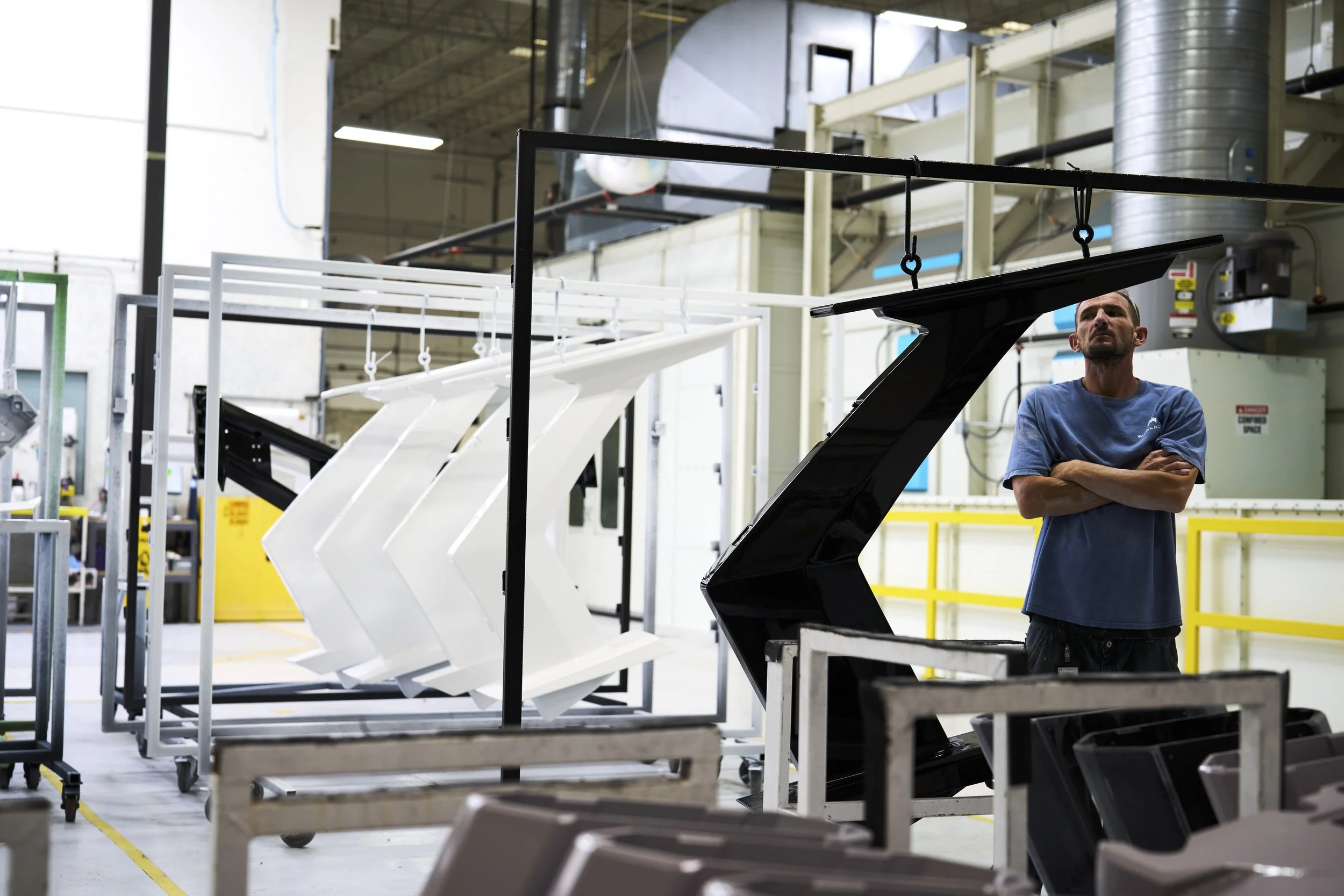 A man in a blue t-shirt stands with arms crossed in a manufacturing or assembly facility, surrounded by large, black and white metal components, machinery, and equipment.