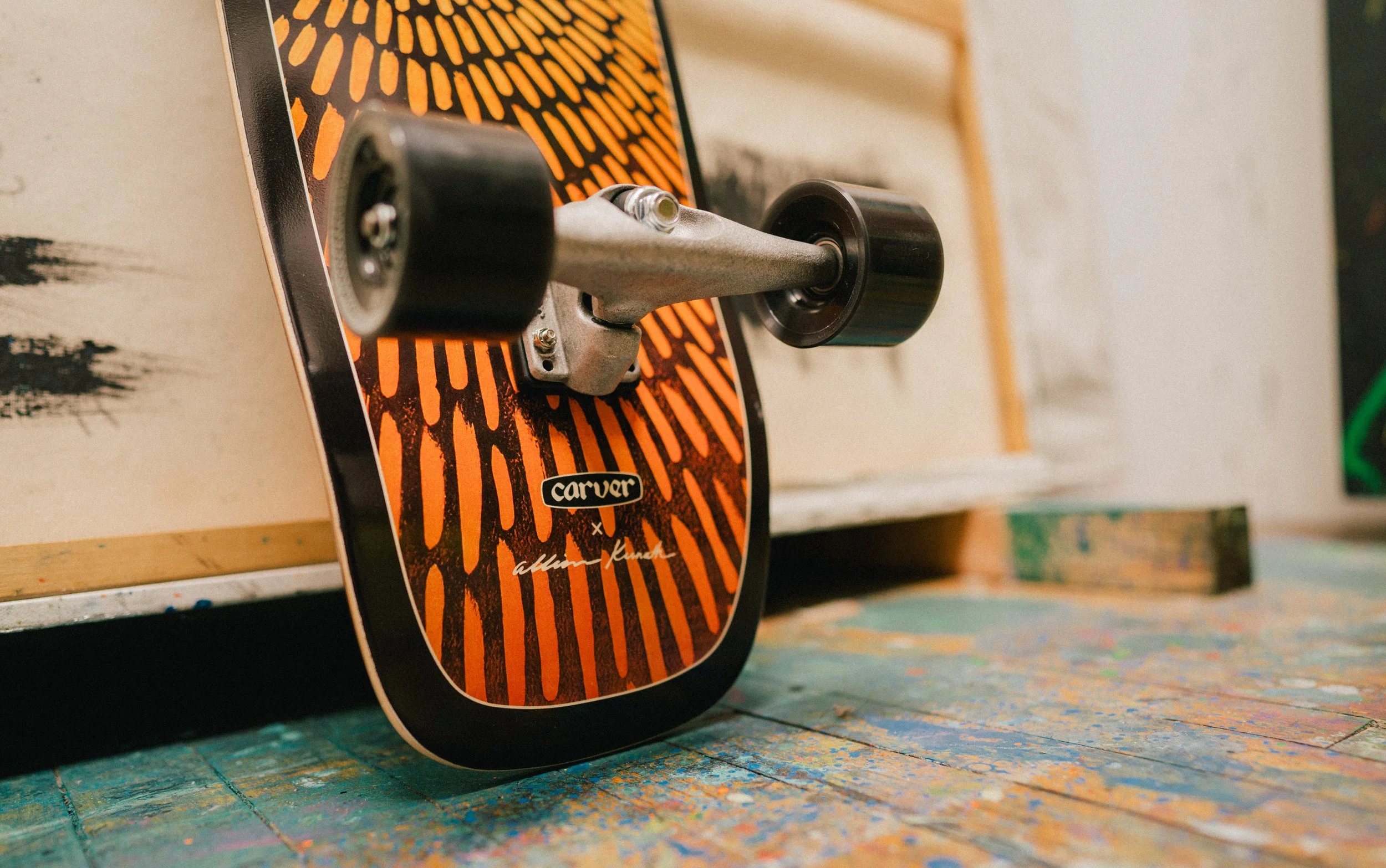 Close-up of a Carver skateboard deck with orange and black design, showing wheels and truck, resting on a colorful painted surface.