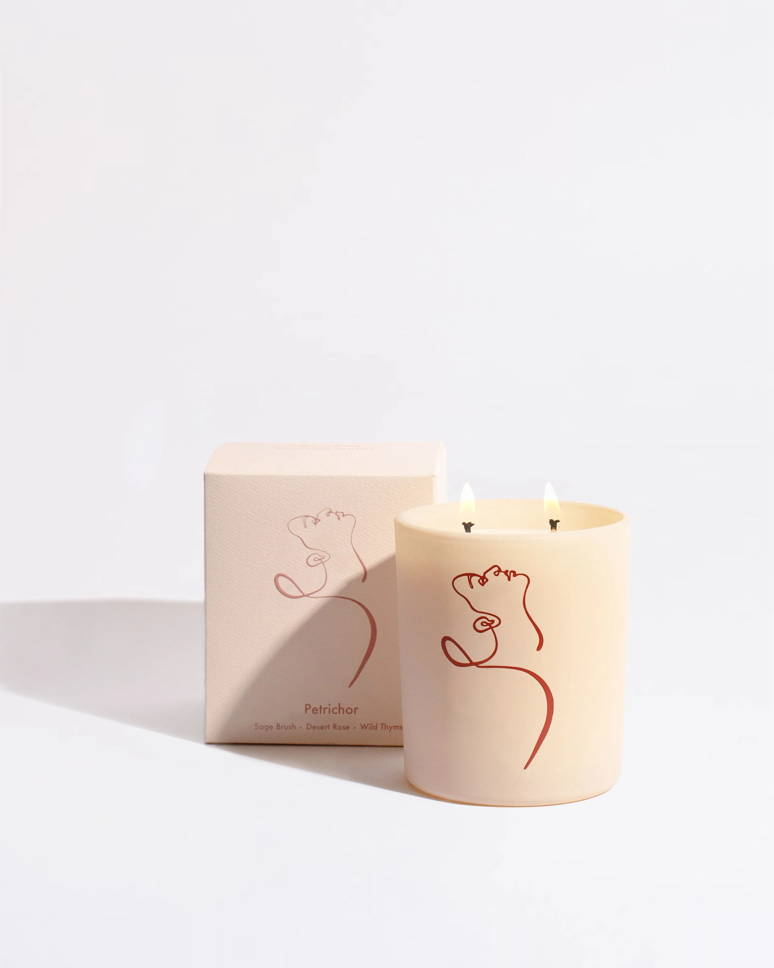 A lit candle with a minimalist line drawing on its surface, alongside a matching box. The design features an abstract profile of a face and the text 'Petrichor' with scent notes: Sage Brush, Desert Rose, Wild Thyme.
