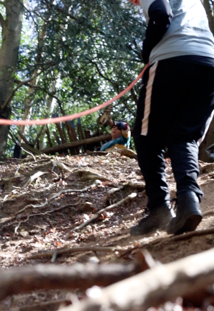 Children hiking in a wooded area with visible roots and dirt, one child is standing and the other is sitting among trees.