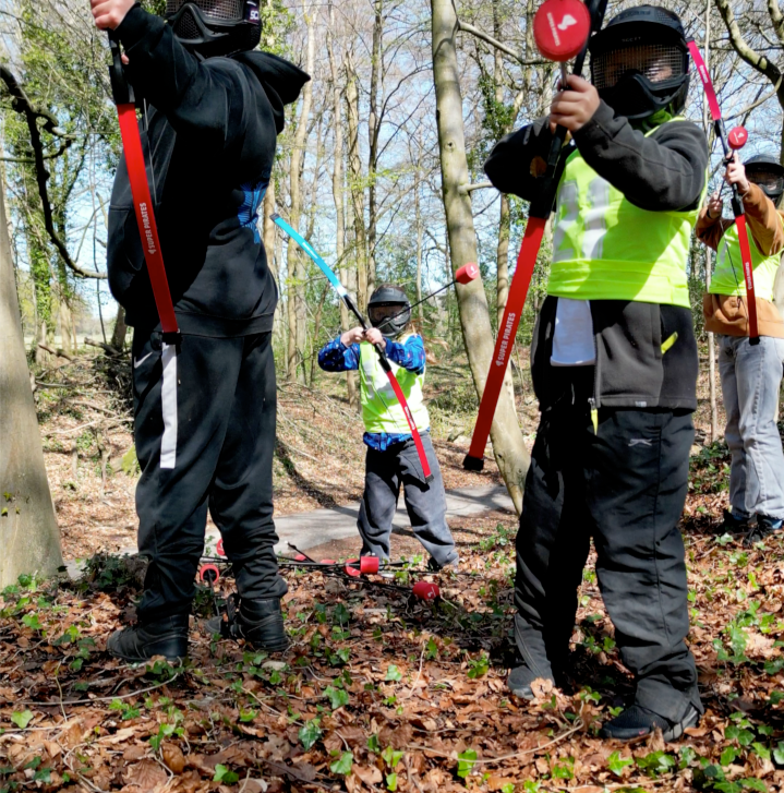 Children playing with toy swords in a wooded park, wearing protective goggles and colorful clothing.