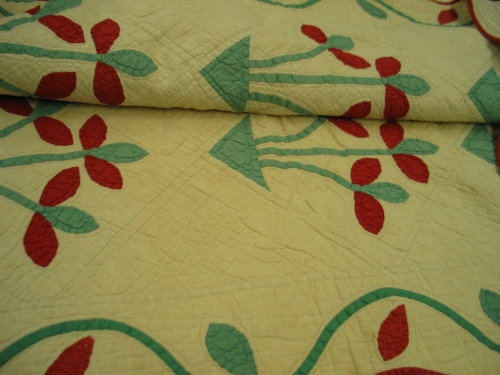 This early 20th-century quilt was hand-sewn with scalloped borders. Its cream-colored background is outlined with red piping. The quilt is filled with green and red flowers.
