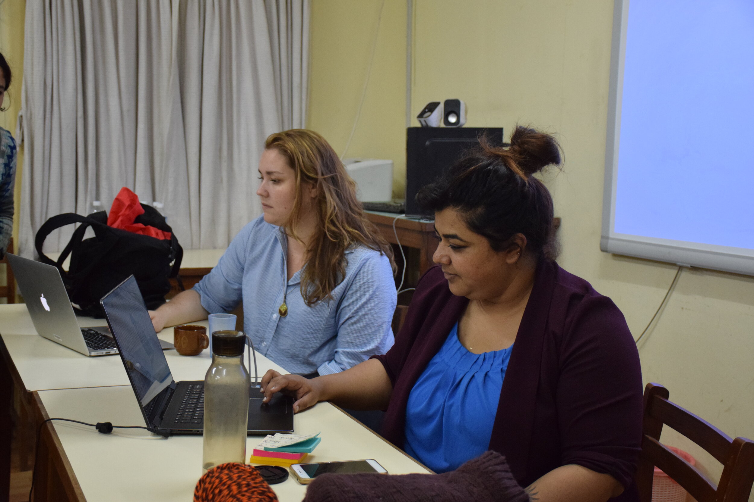 Two workshop facilitators sit in front of a classroom looking down at their computers. 