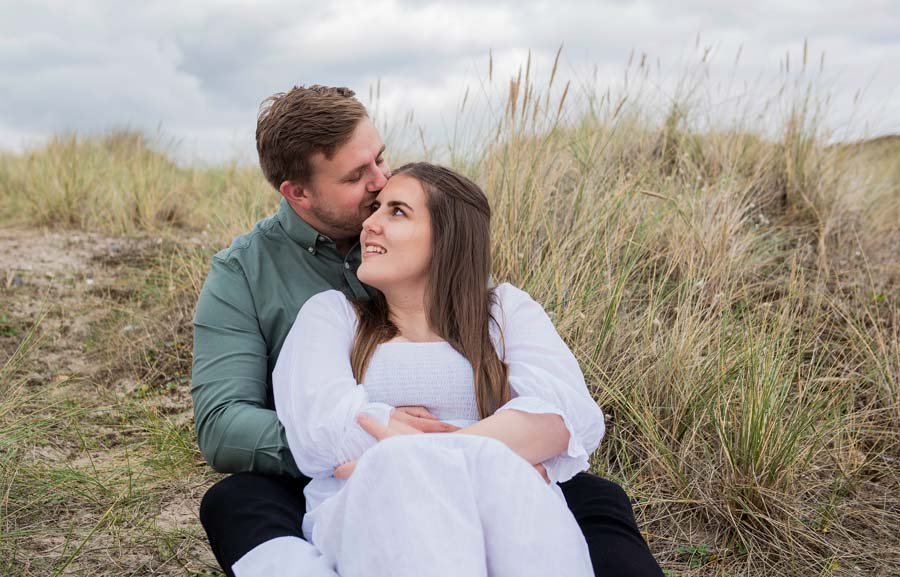 Engaged couple sitting on the sand at West Beach, Littlehampton, as he gently kisses her forehead.