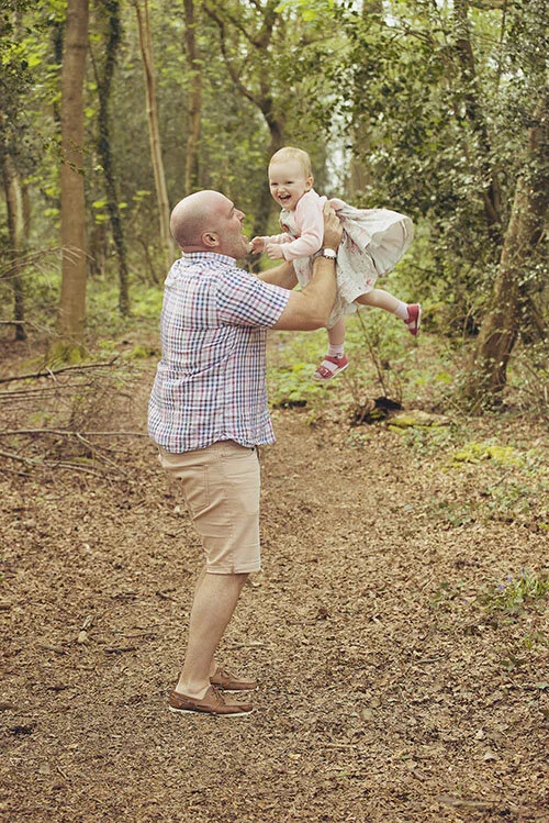 A father lifts his young daughter high into the air in a woodland setting, as she laughs with delight.