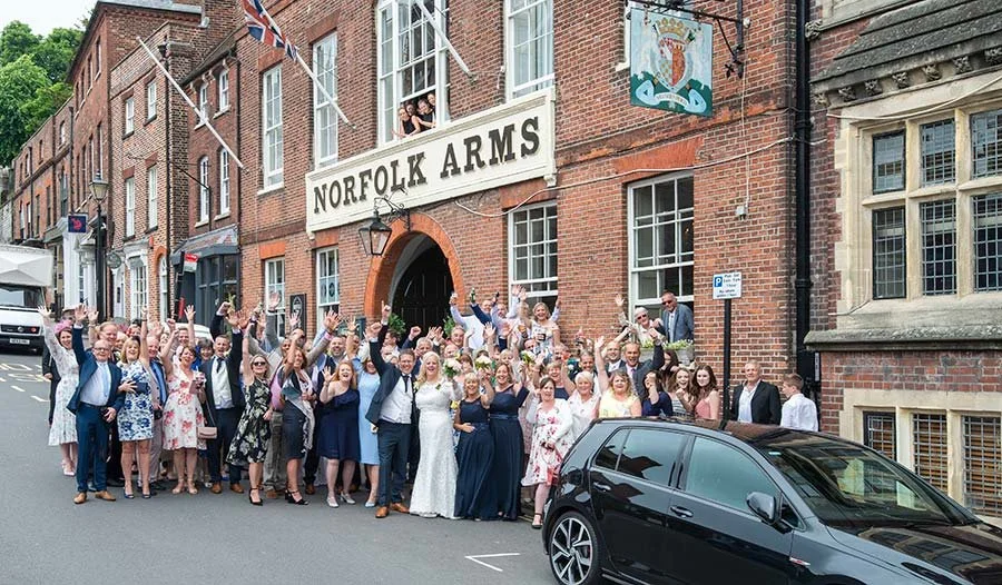 Wedding group photo outside The Norfolk Arms in Arundel, West Sussex, with guests cheering and celebrating around the bride and groom.