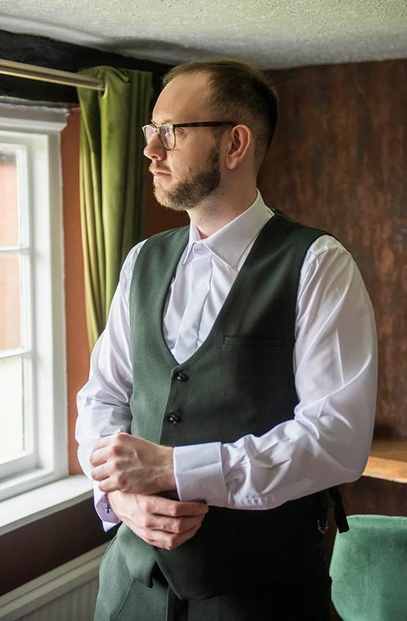 Stylish groom standing by window in suit during morning wedding prep lighting.