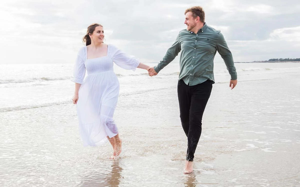 Engaged couple running hand in hand along the sandy beach at West Beach, Littlehampton, smiling towards the camera.