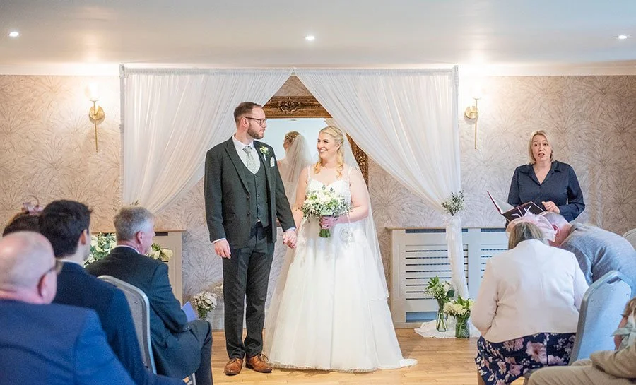 Romantic indoor wedding ceremony at Tottington Manor with floral arch and couple smiling at each other..