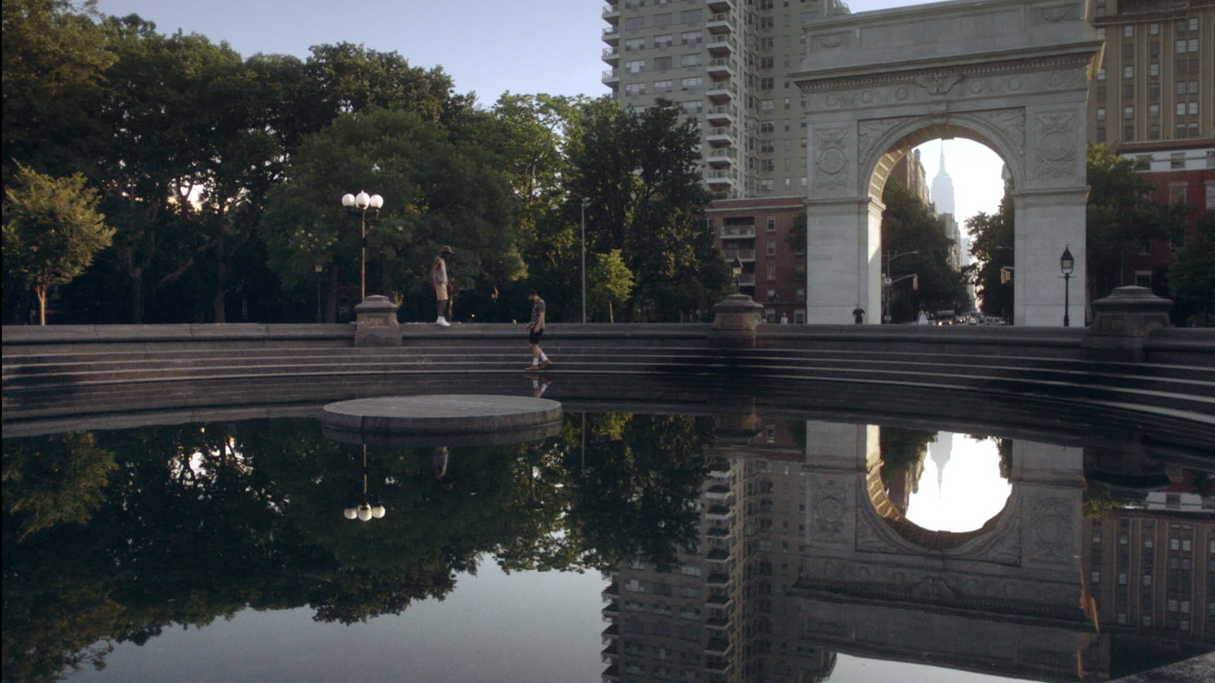 A city park with a stone arch and a fountain, reflecting trees and buildings in the water, with two people near the fountain and high-rise buildings in the background.