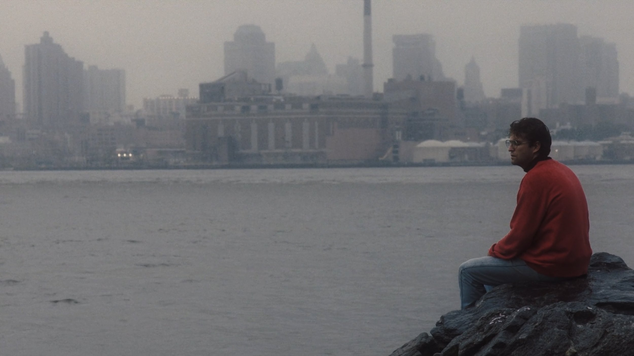 A man in a red jacket sitting on rocks by a waterfront, with a hazy city skyline in the background.