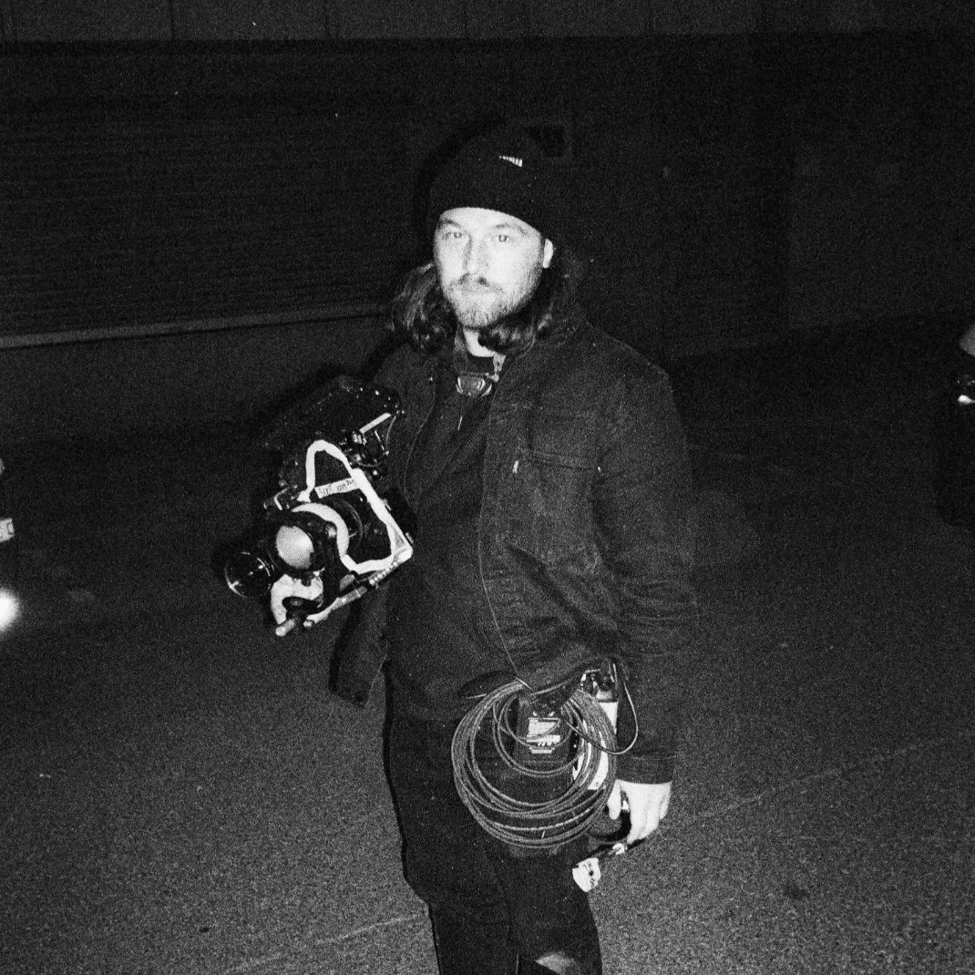 A person with long hair wearing a black beanie and jacket, holding a camera and video equipment, standing outdoors at night.