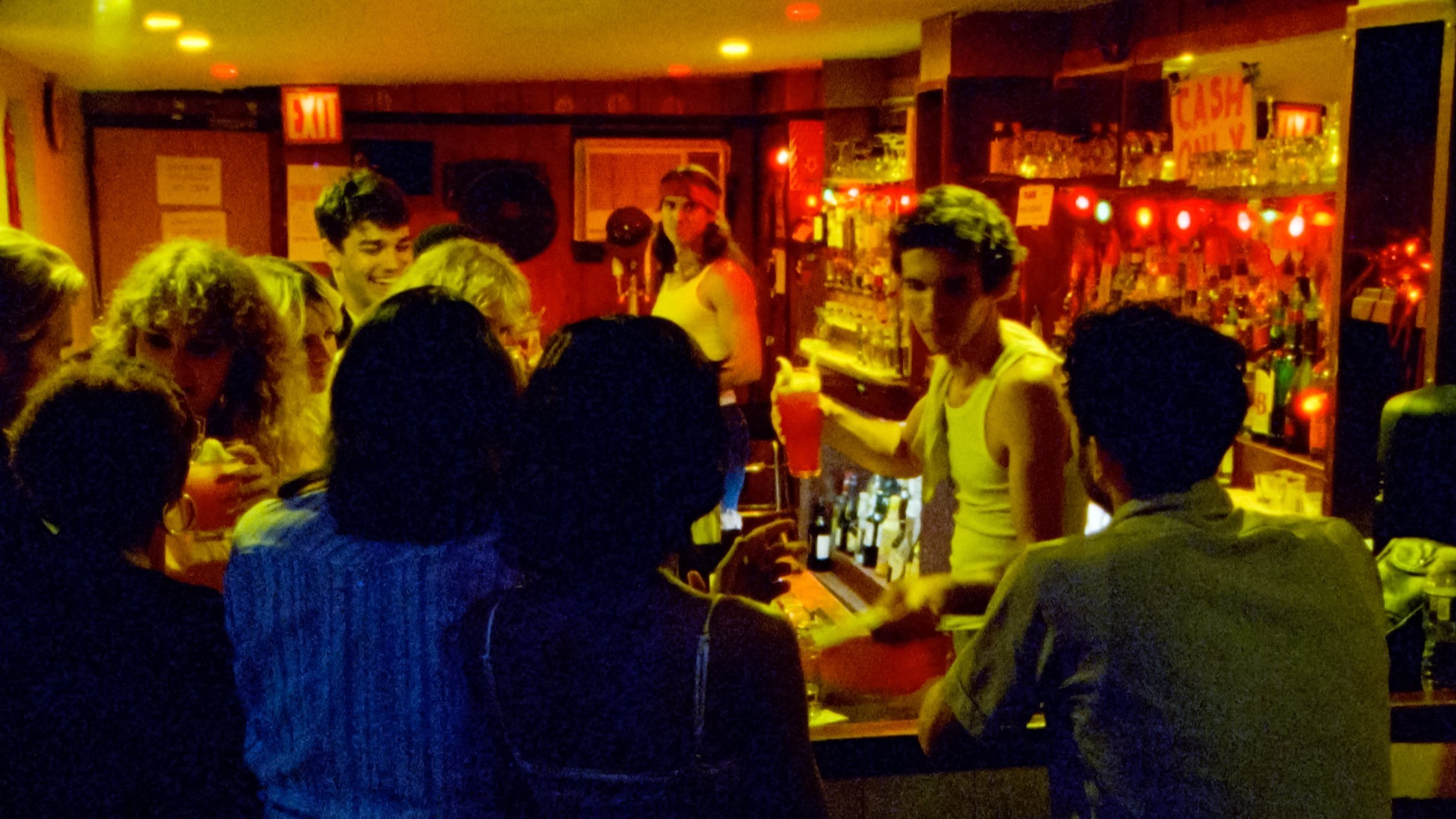 A group of people gathered around a bar in a dimly lit bar or nightclub. The bartender is serving drinks, and there are shelves with bottles and glasses illuminated with red lights.
