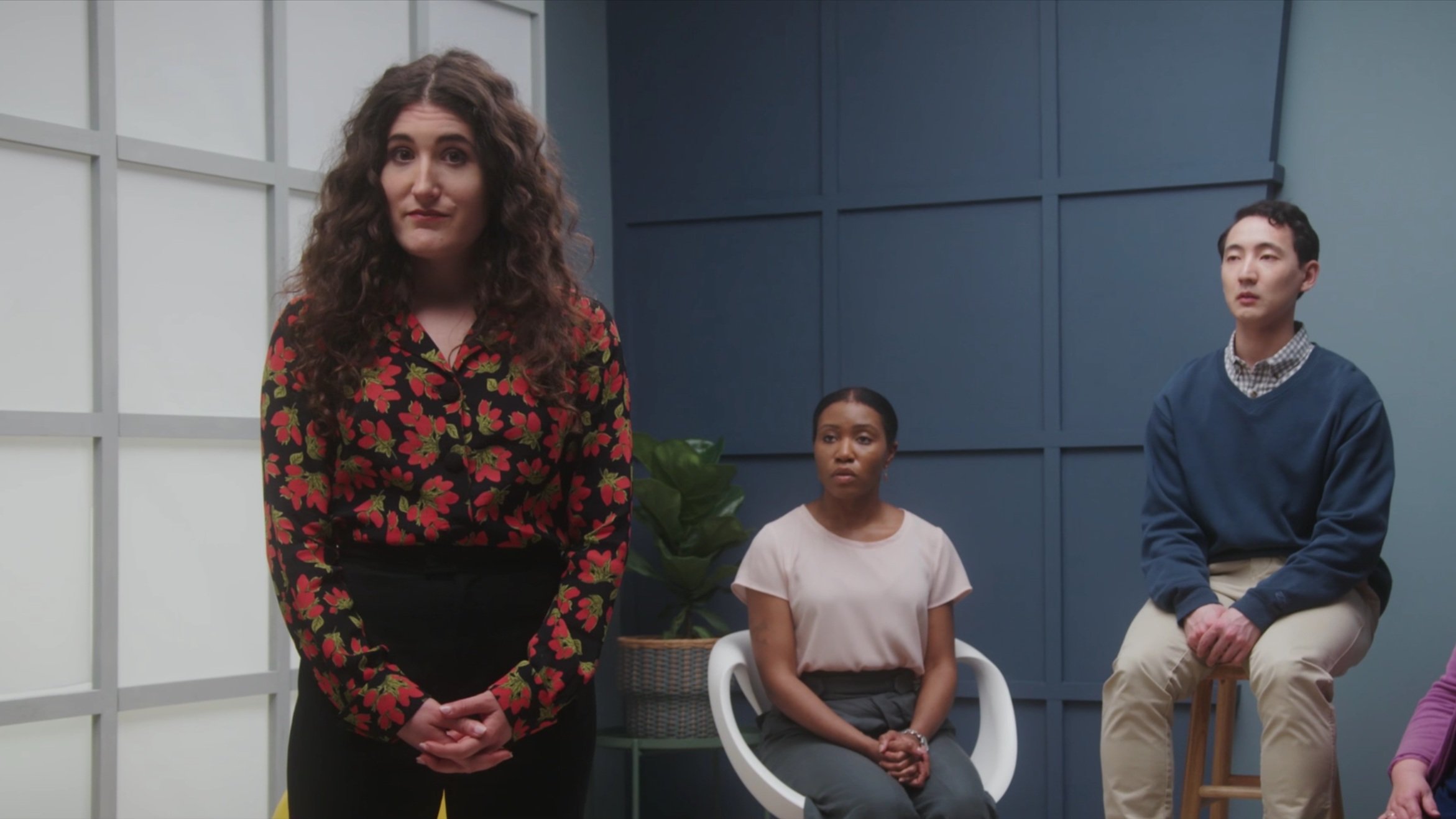 Three people in a meeting room, one woman standing, two seated, with blue walls and a potted plant in the background.