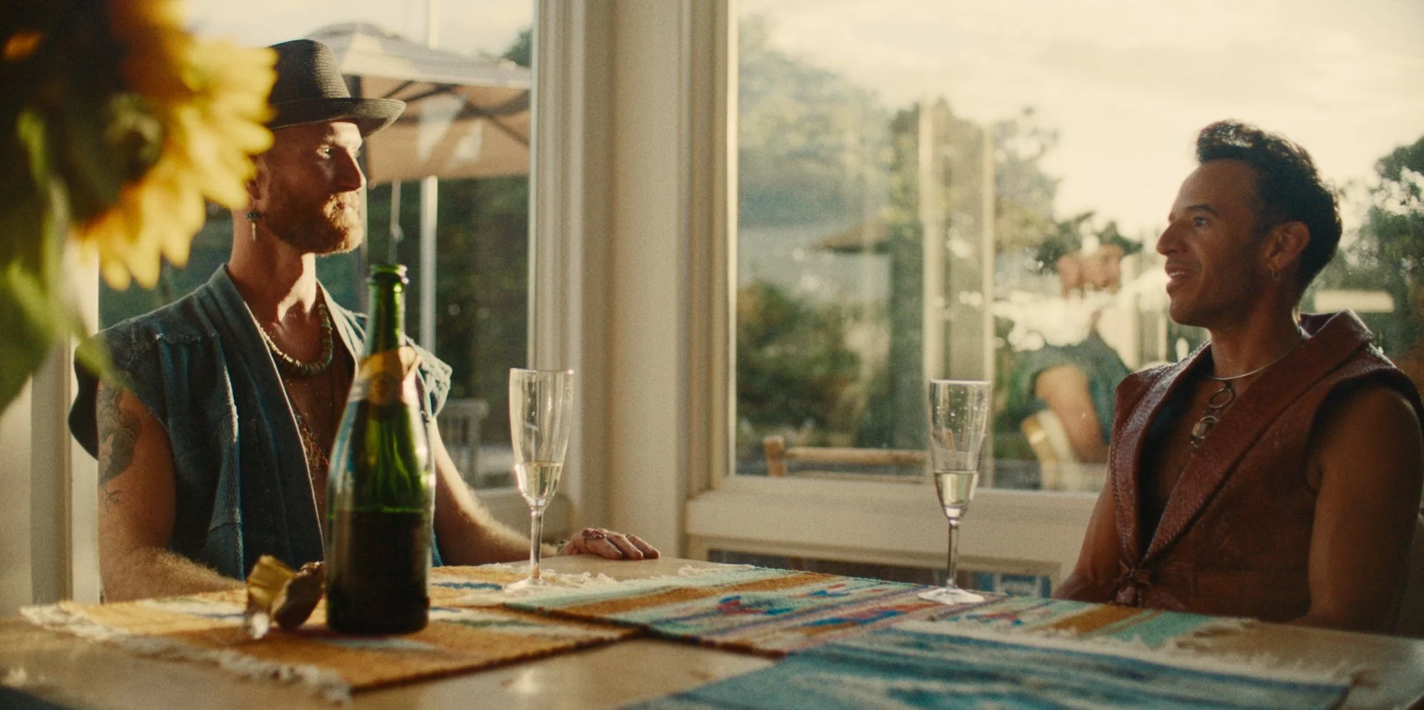 Two men sitting at a dining table on a porch, having a conversation, with sunlight streaming in through large windows. One man wears a hat and a sleeveless denim vest, holding a beer bottle, while the other man has short dark hair, earrings, and a sl